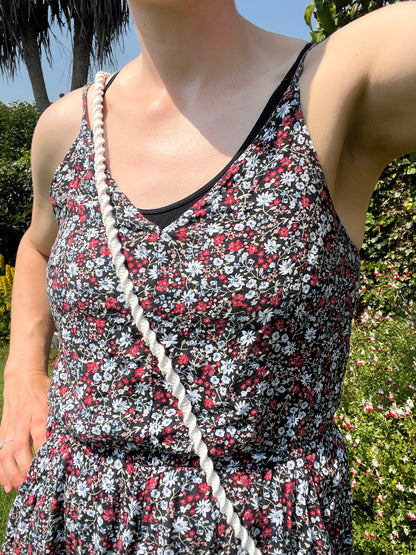 A person wearing a black floral dress and a white braided strap, crafted from recycled cotton cord, is standing outdoors in a garden under bright sunlight. The lush green foliage and colorful flowers form a vibrant backdrop, offering an eco-friendly ambiance. The person's face is out of the frame. They are carrying a Crossbody phone holder (double clip) from Macra-Made-With-Love.