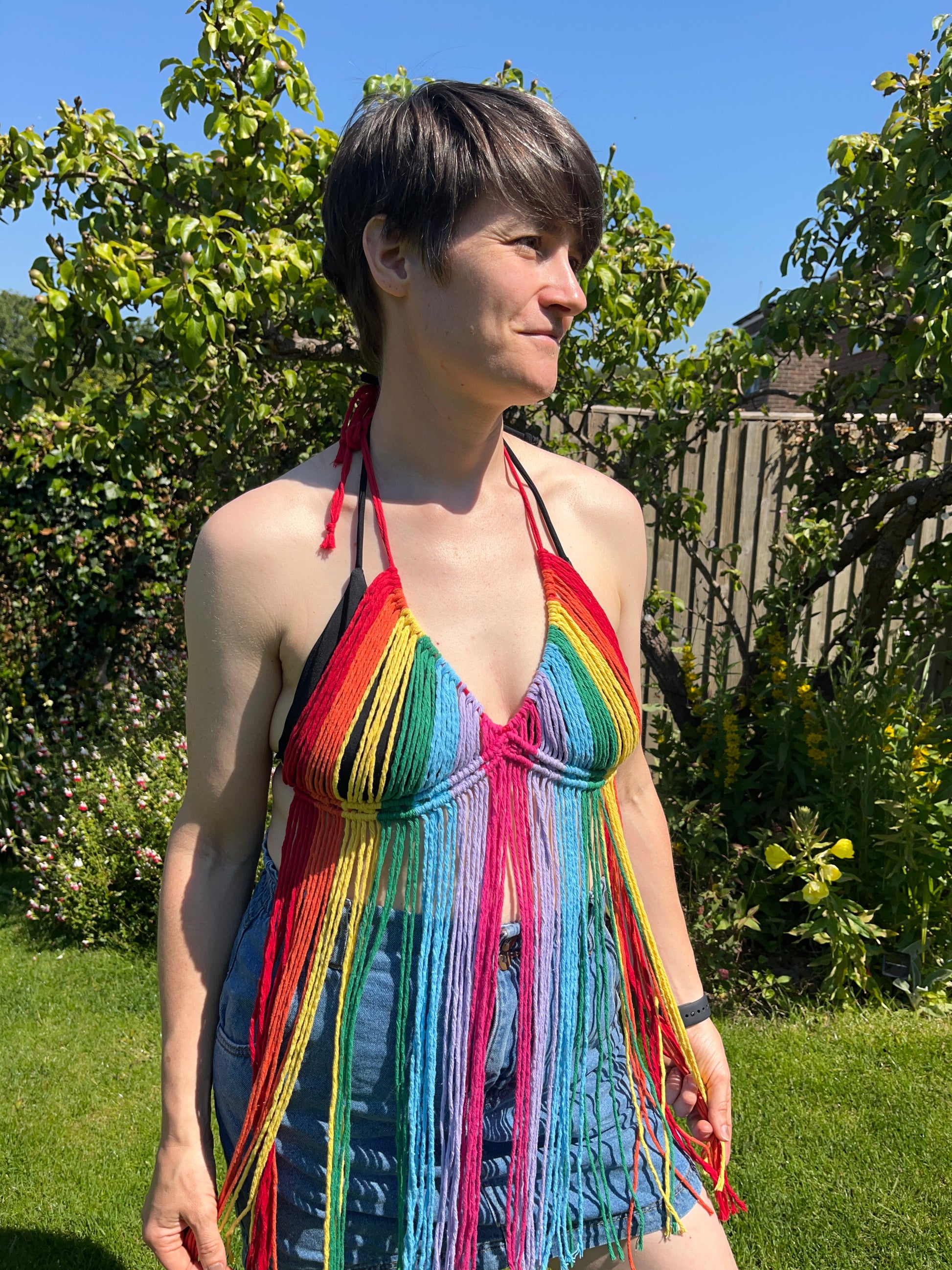 A person stands outdoors on a sunny day, wearing a colorful Rainbow coverup top by Macra-Made-With-Love. They have short dark hair and are looking off to the side. The background features green foliage and a wooden fence, with a bright blue sky overhead—perfect for a festival outfit.