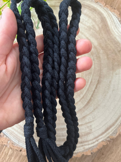 A hand holds multiple strands of thick, twisted black cord, forming a sustainable handmade Braided macrame belt by Macra-Made-With-Love. The background features a textured, circular wooden surface with visible tree rings.