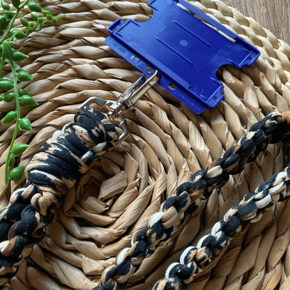 A close-up image of a woven basket tray with an Eco friendly lanyard badge holder - 34” by Macra-Made-With-Love in patterned black, beige, and white. The lanyard features a silver clasp attached to a blue plastic ID badge holder. A small green succulent is placed in the upper left corner.