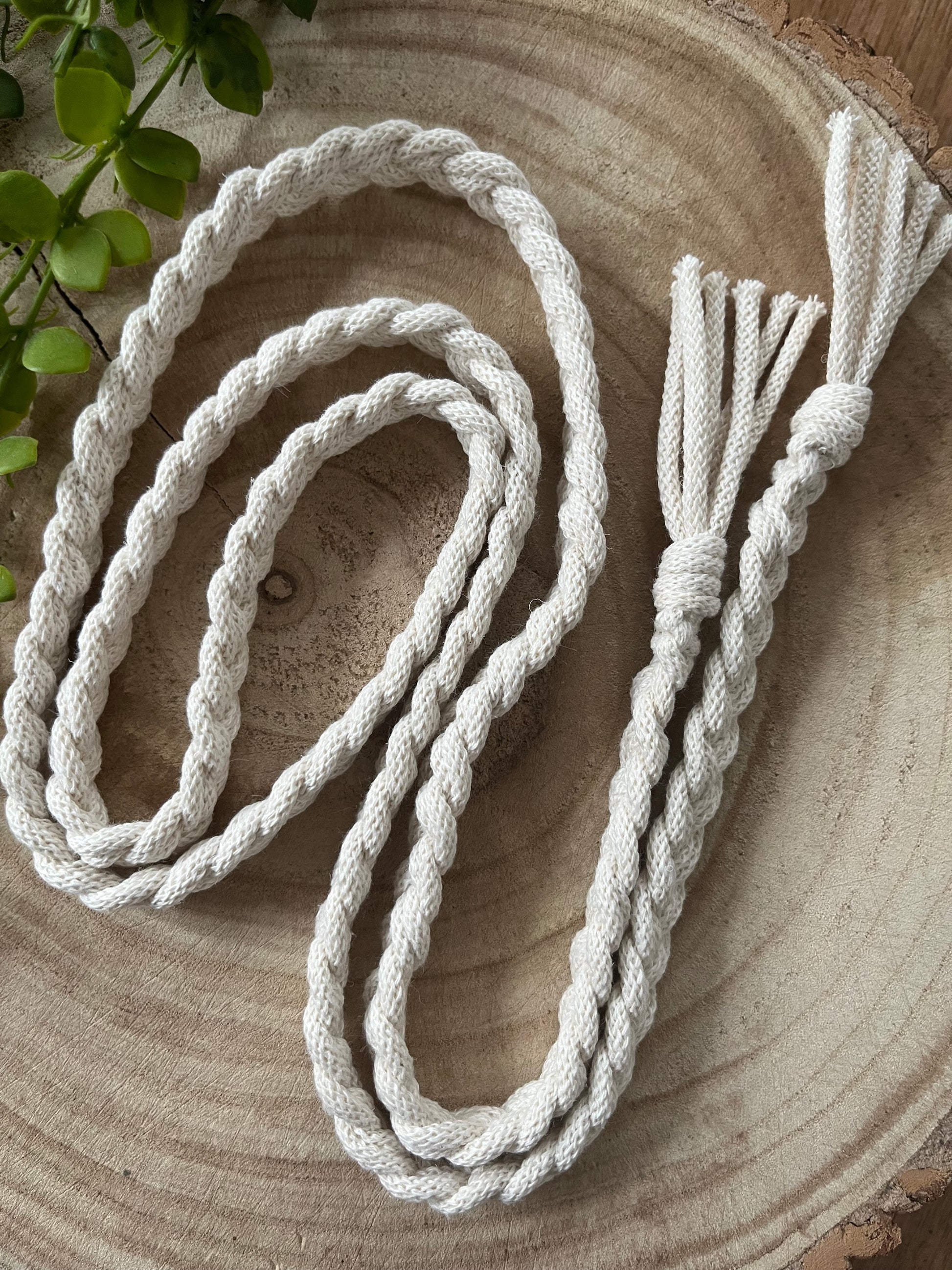 A neatly coiled, thick, white **Braided macrame belt** by **Macra-Made-With-Love** made from recycled cotton braided yarn is placed on a round wooden surface. The belt's end features a handmade knot belt section with fringed ends. A small green plant is partially visible at the top left corner.