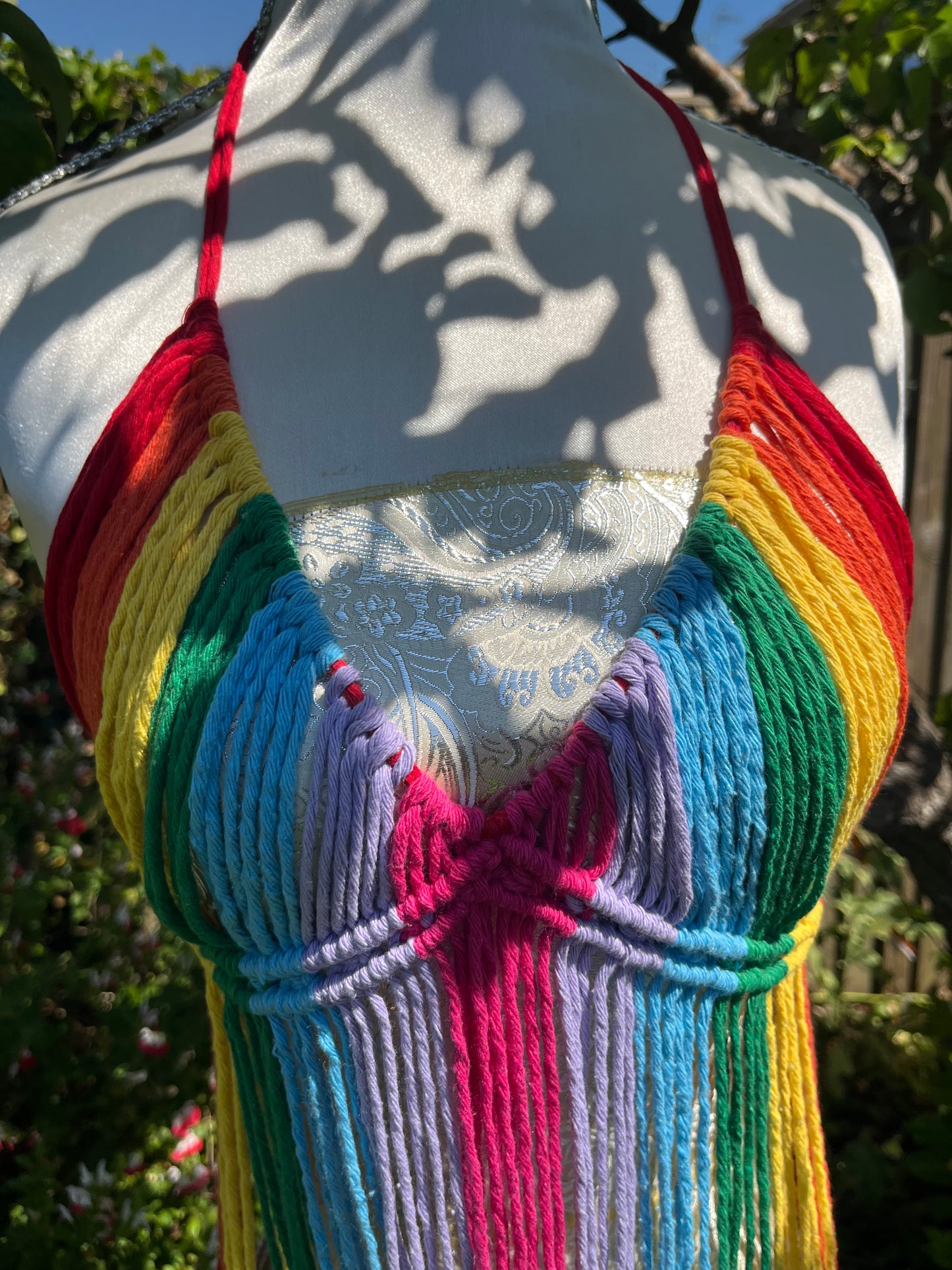 A mannequin wearing a colorful, intricately woven festival outfit with a halter neckline displayed outdoors. The Rainbow coverup top by Macra-Made-With-Love features vibrant, textured yarn in red, yellow, green, blue, purple, and pink, with a detailed floral pattern on the white section underneath.