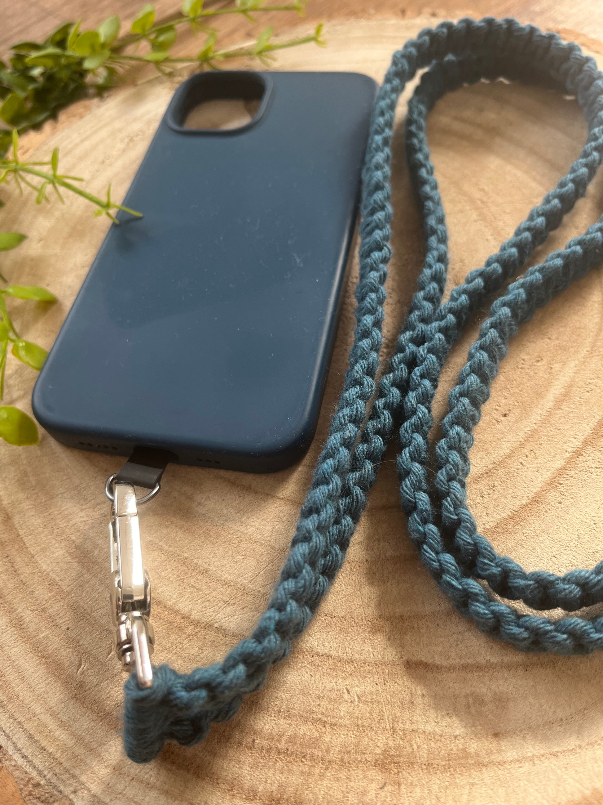 A blue phone case with an attached braided strap is placed on a wooden surface. Crafted from eco-friendly, recycled cotton, the Macra-Made-With-Love Recycled Cotton Adjustable Crossbody Phone Strap with Universal Tether adds both style and sustainability. Greenery is visible in the background.