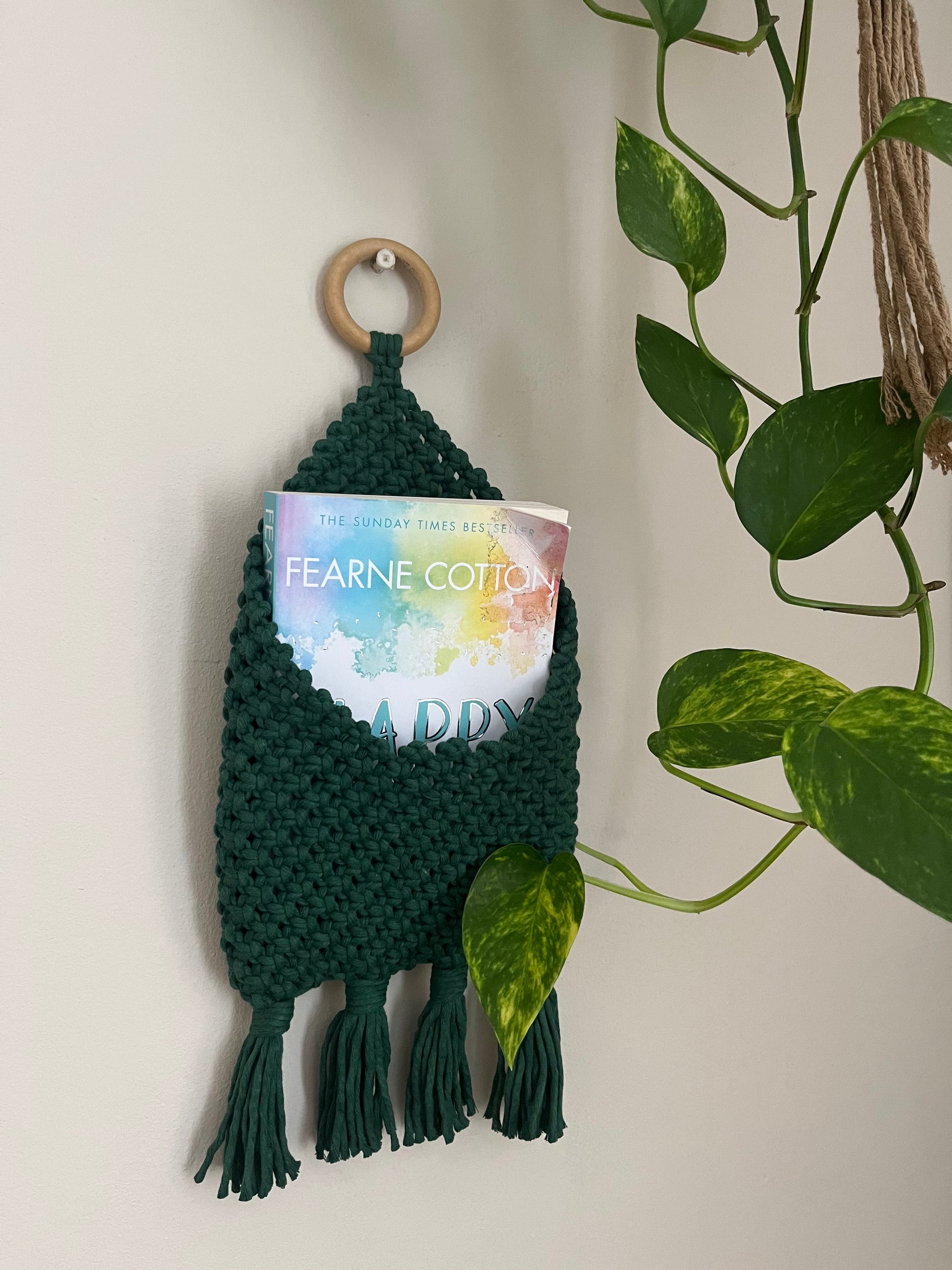 A Bedside storage pouch book sleeve by Macra-Made-With-Love, with braided details and tassels, made from recycled cotton decor, is holding a book titled "Happy" by Fearne Cotton. This eco-friendly bedroom storage solution is attached to a wooden ring and displayed on a white wall next to a leafy green plant.