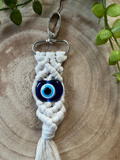 An Evil eye keychain from Macra-Made-With-Love with a blue evil eye bead in the center is displayed on a circular wooden surface. This protection talisman is made from white braided cord and has a silver clasp at the top. Green leaves can be seen in the background.