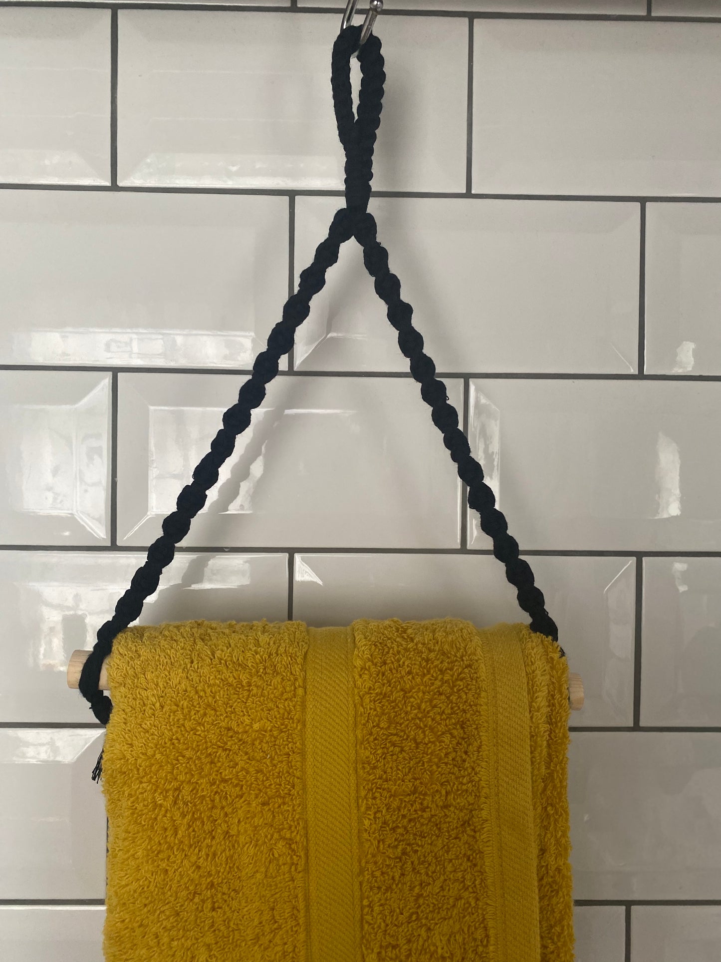 A thick, yellow towel made from sustainable recycled cotton hangs on a Macra-Made-With-Love Hand towel holder against a white tiled wall. The eco-friendly rope is looped around a metal hook at the top, with the ends attached to a wooden rod on which the towel is draped.