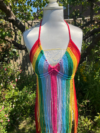 A dress form outdoors displays a colorful, fringed Rainbow coverup top with horizontal rainbow stripes. This bohemian Macra-Made-With-Love top stands out against the background of greenery, flowers, and a wooden fence, suggesting a garden setting—perfect for a festival outfit.