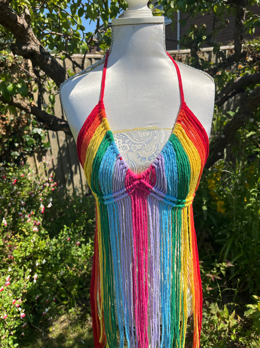 A dress form outdoors displays a colorful, fringed Rainbow coverup top with horizontal rainbow stripes. This bohemian Macra-Made-With-Love top stands out against the background of greenery, flowers, and a wooden fence, suggesting a garden setting—perfect for a festival outfit.