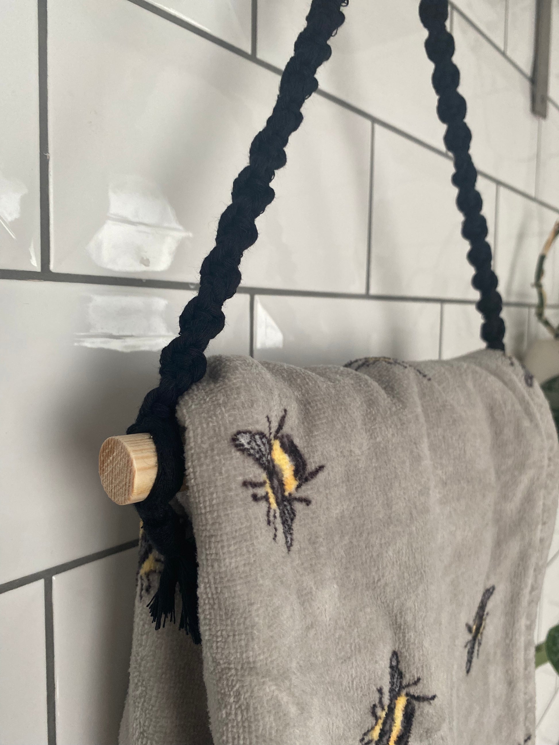 A gray towel made from sustainable recycled cotton, adorned with embroidered bees, hangs on a black braided rope looped around a wooden dowel. The eco-friendly towel is displayed against a white tiled wall, elegantly held by the Hand Towel Holder from Macra-Made-With-Love.