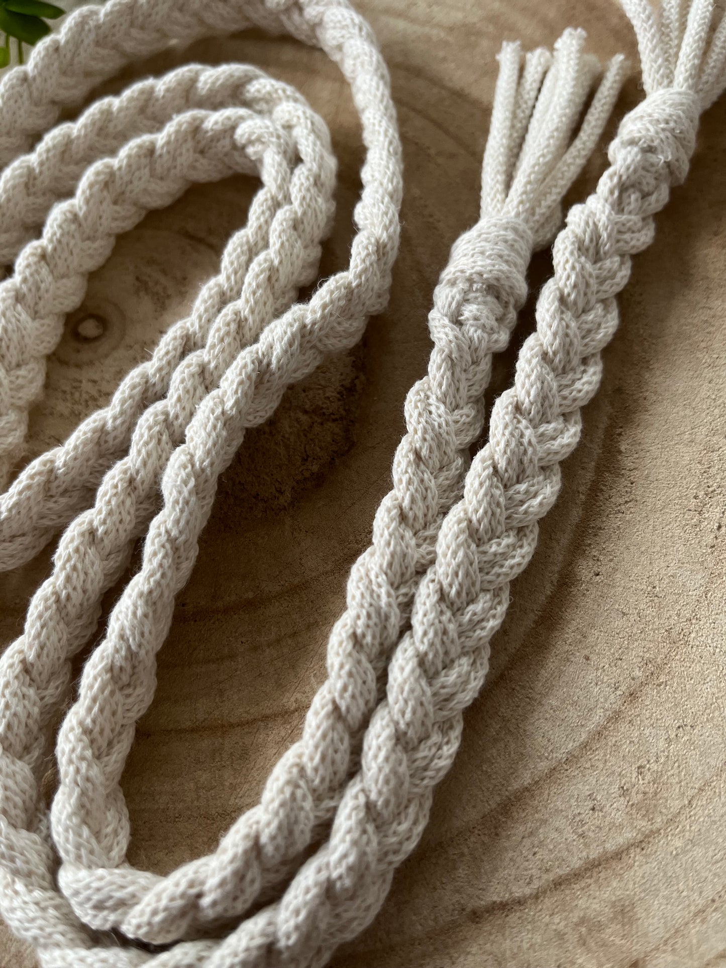 Close-up of intricately braided white cotton ropes resting on a wooden surface. The textures of the tightly woven strands and the wood's natural grain provide a rustic yet neat appearance, much like an eco-friendly waist accessory. The Braided macrame belt by Macra-Made-With-Love features ends that are neatly frayed, adding a handmade charm.