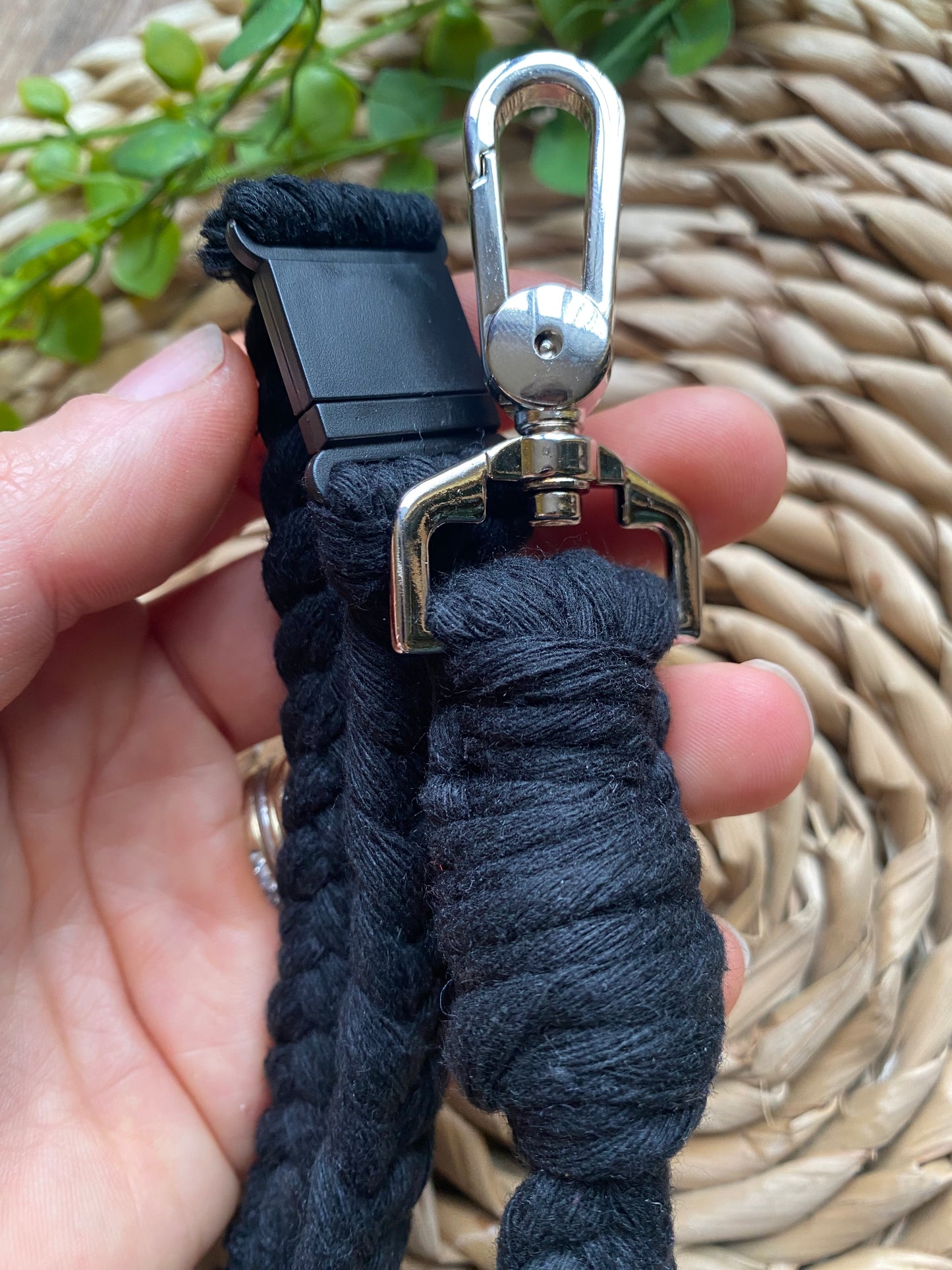 Close-up of a hand holding an Eco friendly lanyard badge holder - 34” from Macra-Made-With-Love, featuring a metal clip at the end for attachment. The background shows part of a woven basket and some green plant leaves.