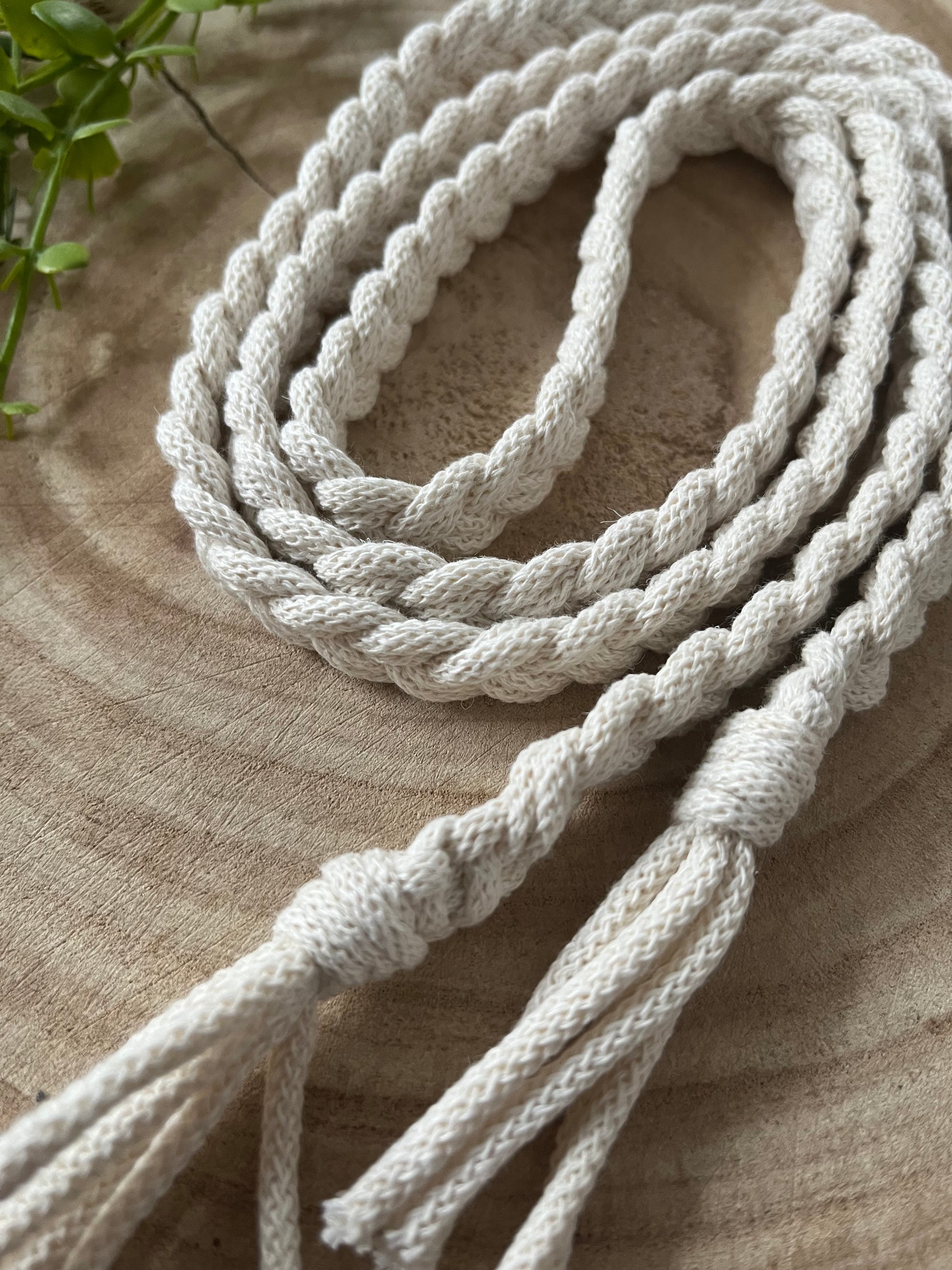 Close-up of a coiled, twisted white rope with frayed ends, resting on a wooden surface. Green leaves are visible in the background. The rope's textured appearance suggests it could be an **Eco friendly braided macrame belt - waist accessory** crafted from natural fibers by **Macra-Made-With-Love**.