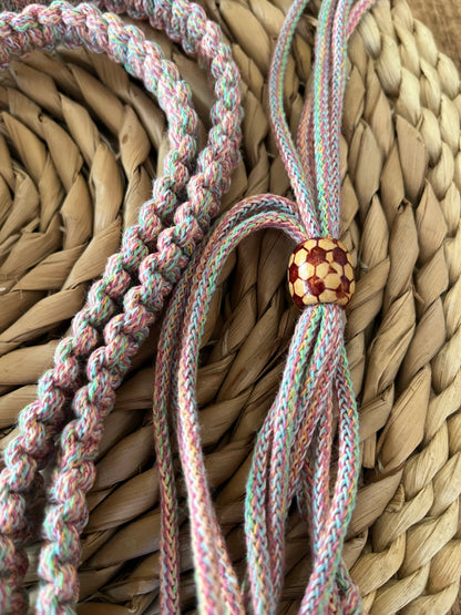 Close-up of colorful braided threads in various shades of pink, blue, and yellow arranged on a woven straw surface. One set of threads features a round bead adorned with a red and yellow mosaic pattern. This Macra-Made-With-Love Recycled Cotton Adjustable Crossbody Phone Strap with Universal Tether is an eco-friendly phone accessory made from recycled cotton braided cord.