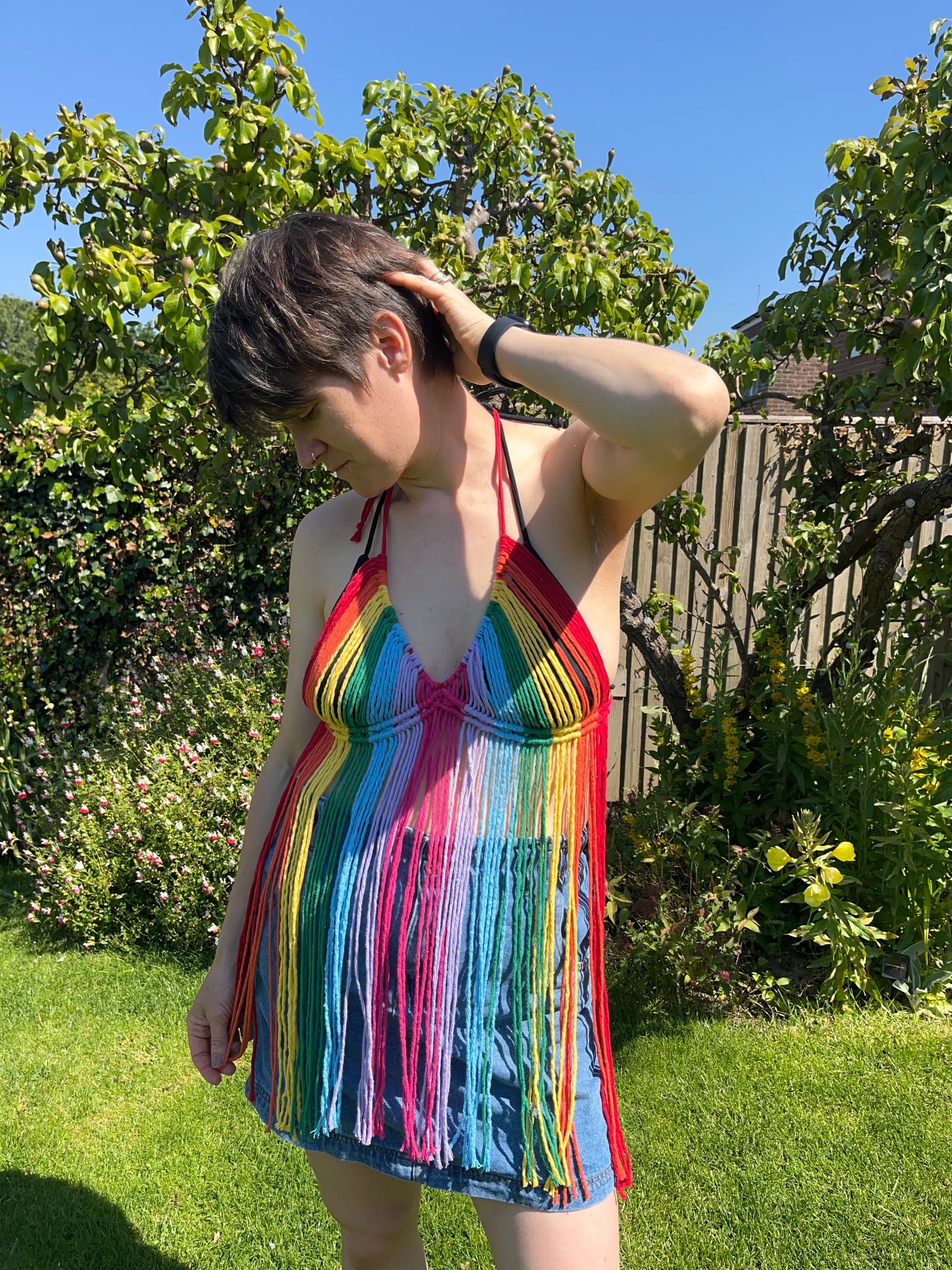 A person with short hair stands in a green garden, wearing a Macra-Made-With-Love Rainbow coverup top over a denim skirt. They are looking down and touching their head with one hand. The background features trees, plants, and a wooden fence.