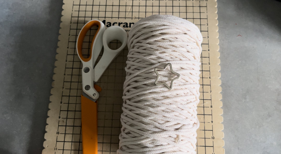 A pair of orange-handled scissors lies beside a spool of white cord on a grid-patterned macrame board. A star-shaped metal ring is attached to the cord, perfect for the Macra-Made-With-Love Macrame Christmas Tree tutorial. The scene is set on a gray surface.