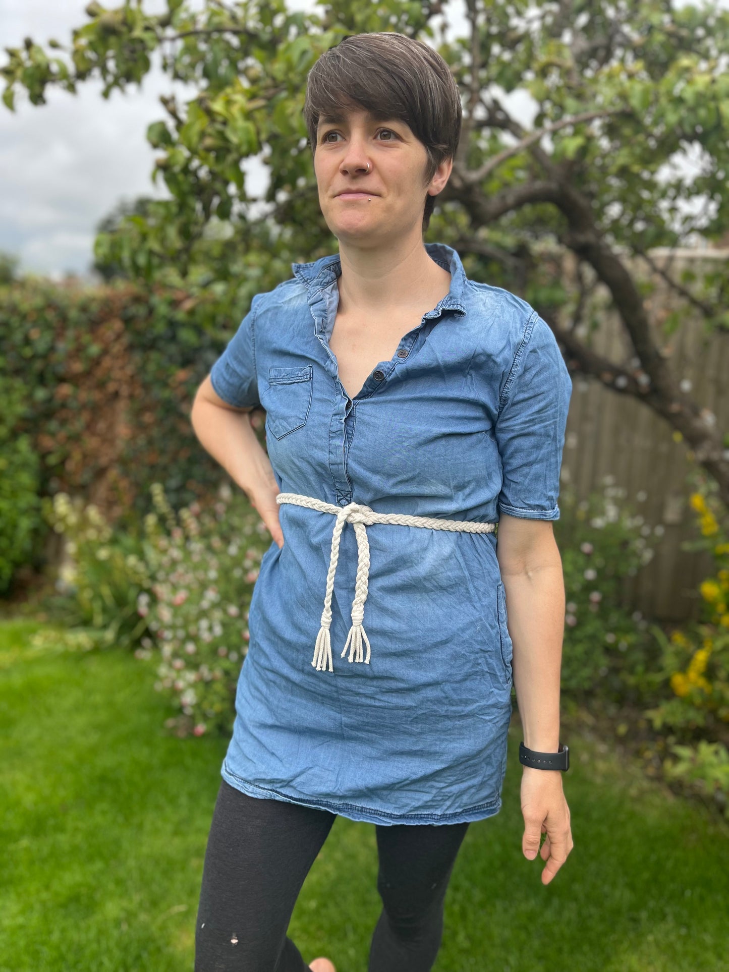 A person stands in a garden with green grass and plants in the background. They are wearing a denim dress with an eco-friendly waist accessory, featuring the Braided Macrame Belt from Macra-Made-With-Love, made from recycled cotton braided yarn and a black fitness watch. Their short brown hair is styled simply, and they appear to be looking off into the distance.