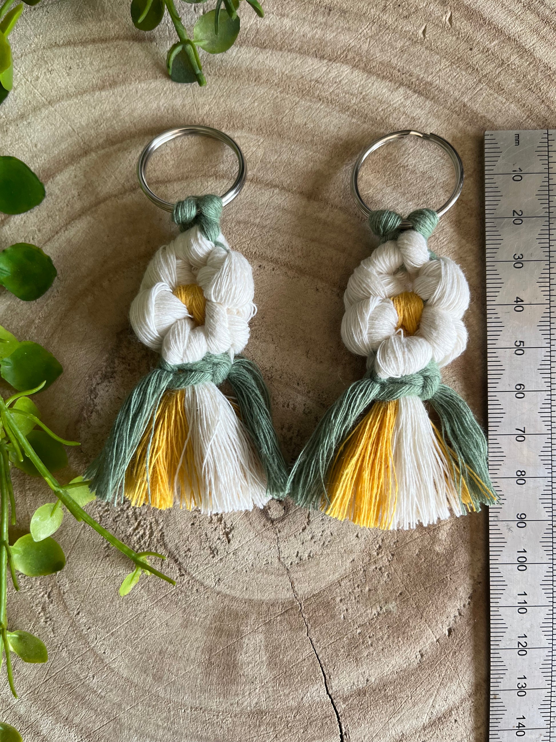 A pair of handmade macrame daisy boot charms, made with recycled cotton, on a wooden surface, next to a metal measuring ruler. 