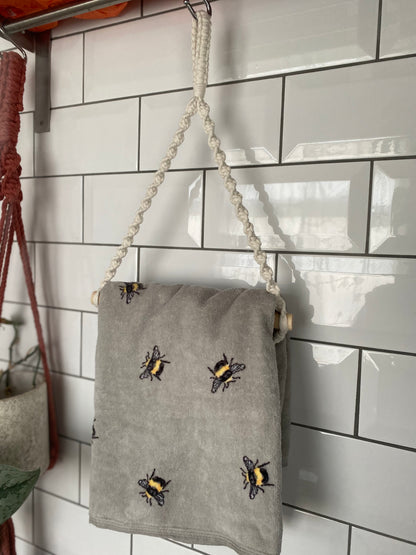 A gray towel made from sustainable recycled cotton, with yellow and black bee designs, hangs on a white tile wall in a kitchen or bathroom. The towel is suspended by a looped, rope hanger attached to a metallic rod. To the left, a plant in a ceramic pot hangs from an eco-friendly macrame holder called the Hand towel holder by Macra-Made-With-Love.