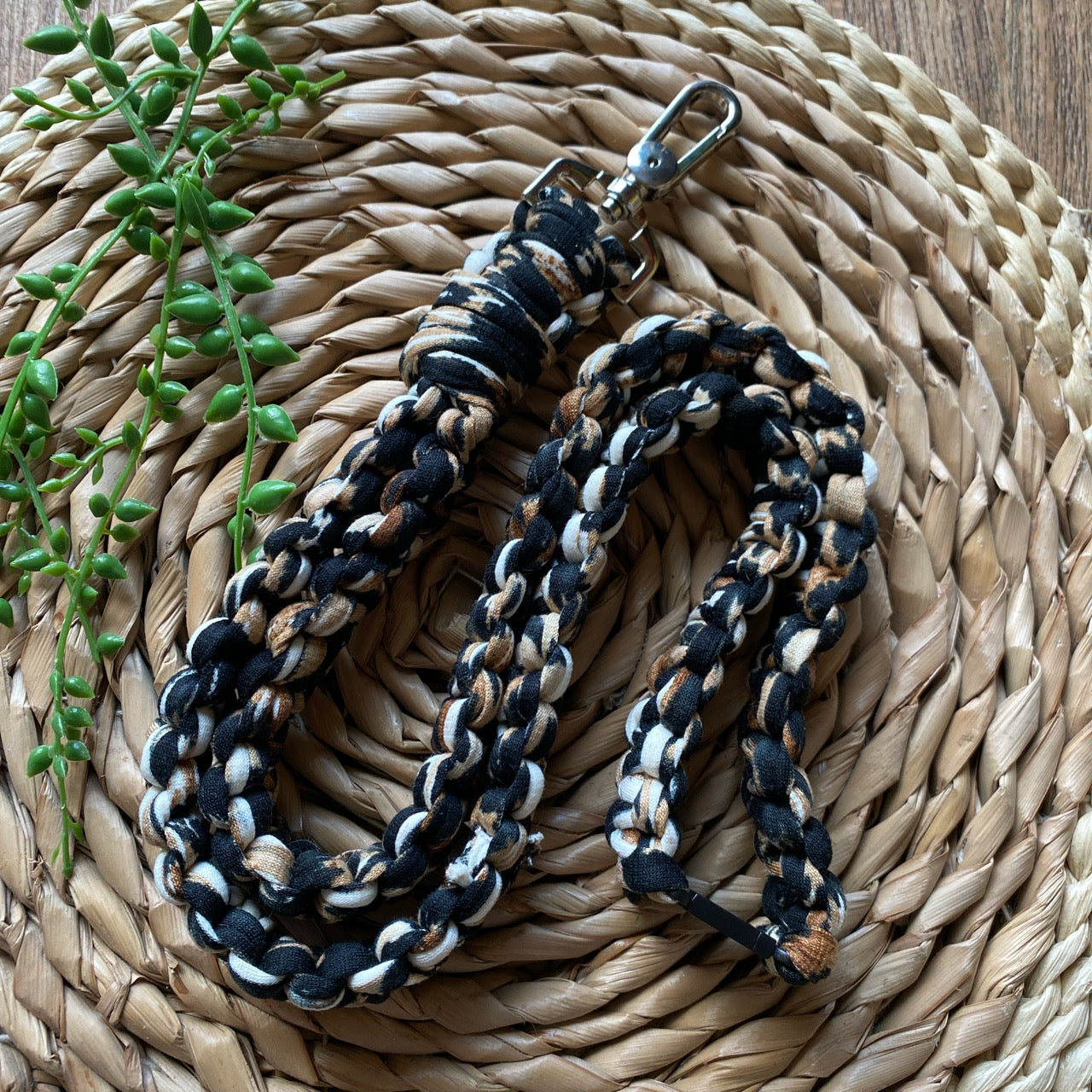 A braided dog leash with black, white, and tan colors and a metal clasp lays on a woven straw mat. A small sprig of greenery is placed beside the Macra-Made-With-Love Eco friendly lanyard badge holder - 34” on the left side of the image.