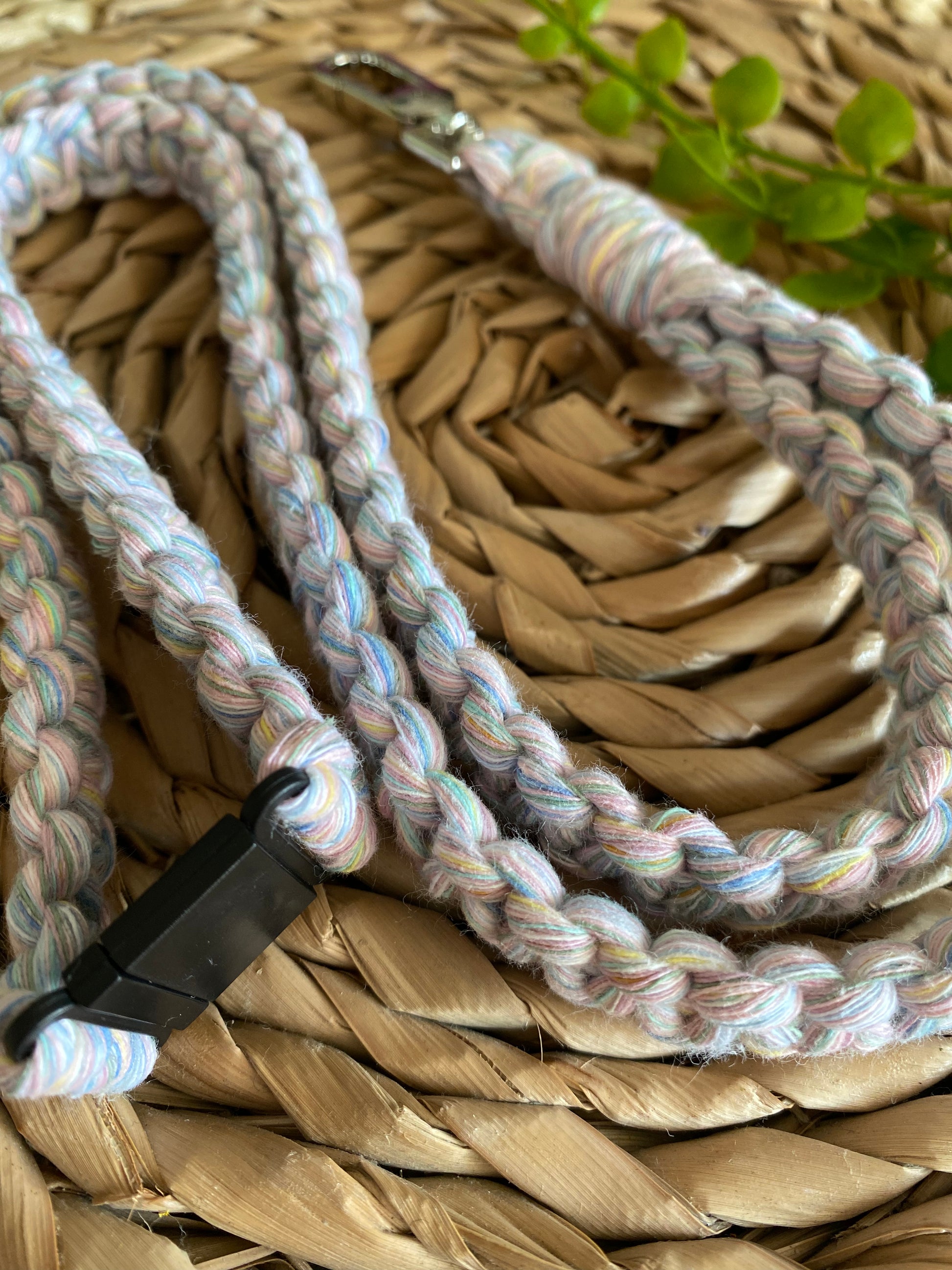 A braided pastel-colored cord with a black plastic clasp, doubling as an Eco friendly lanyard badge holder - 34” by Macra-Made-With-Love, is placed on a woven mat. Small green leaves are present in the background, adding a touch of nature to the scene.