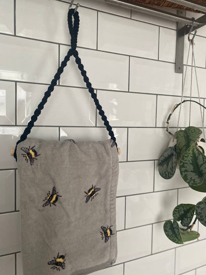 A gray towel made from sustainable recycled cotton and adorned with embroidered bees hangs on a black braided rope against a white tiled wall. To the right, a Macra-Made-With-Love Hand Towel Holder holds a potted plant with heart-shaped leaves. Another towel rack is partially visible above the towel.