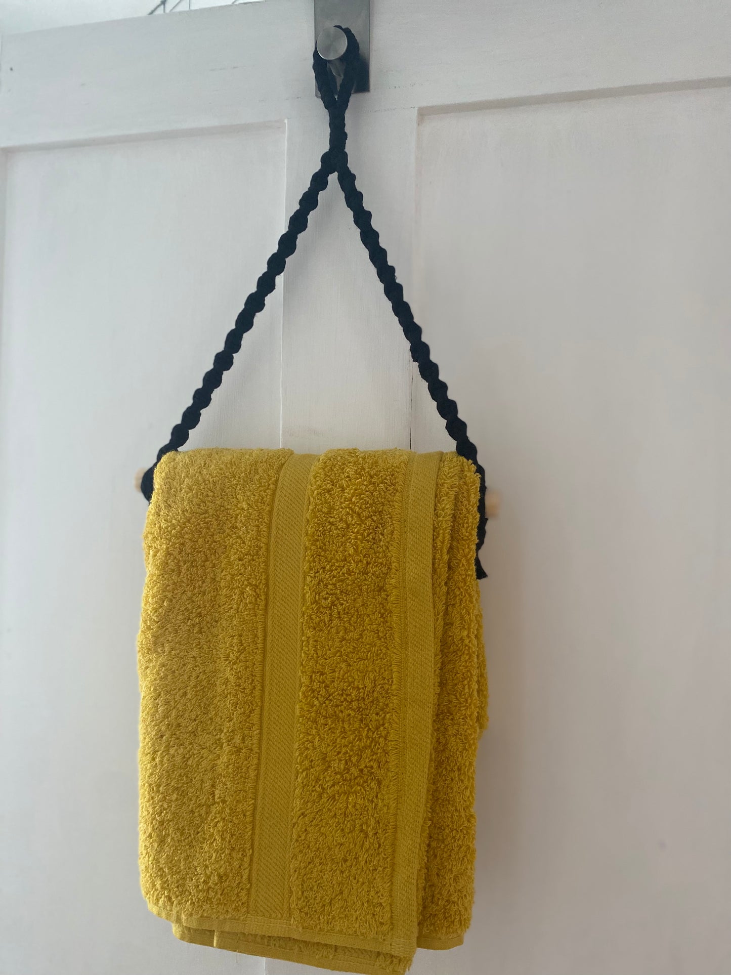 A yellow towel made from sustainable recycled cotton is hanging on a white wooden door by a black braided cord and wooden rod from the Hand towel holder by Macra-Made-With-Love. The towel is neatly folded, with two vertical stripes along its edge. The simple, white-painted door contrasts with the bright eco-friendly towel.