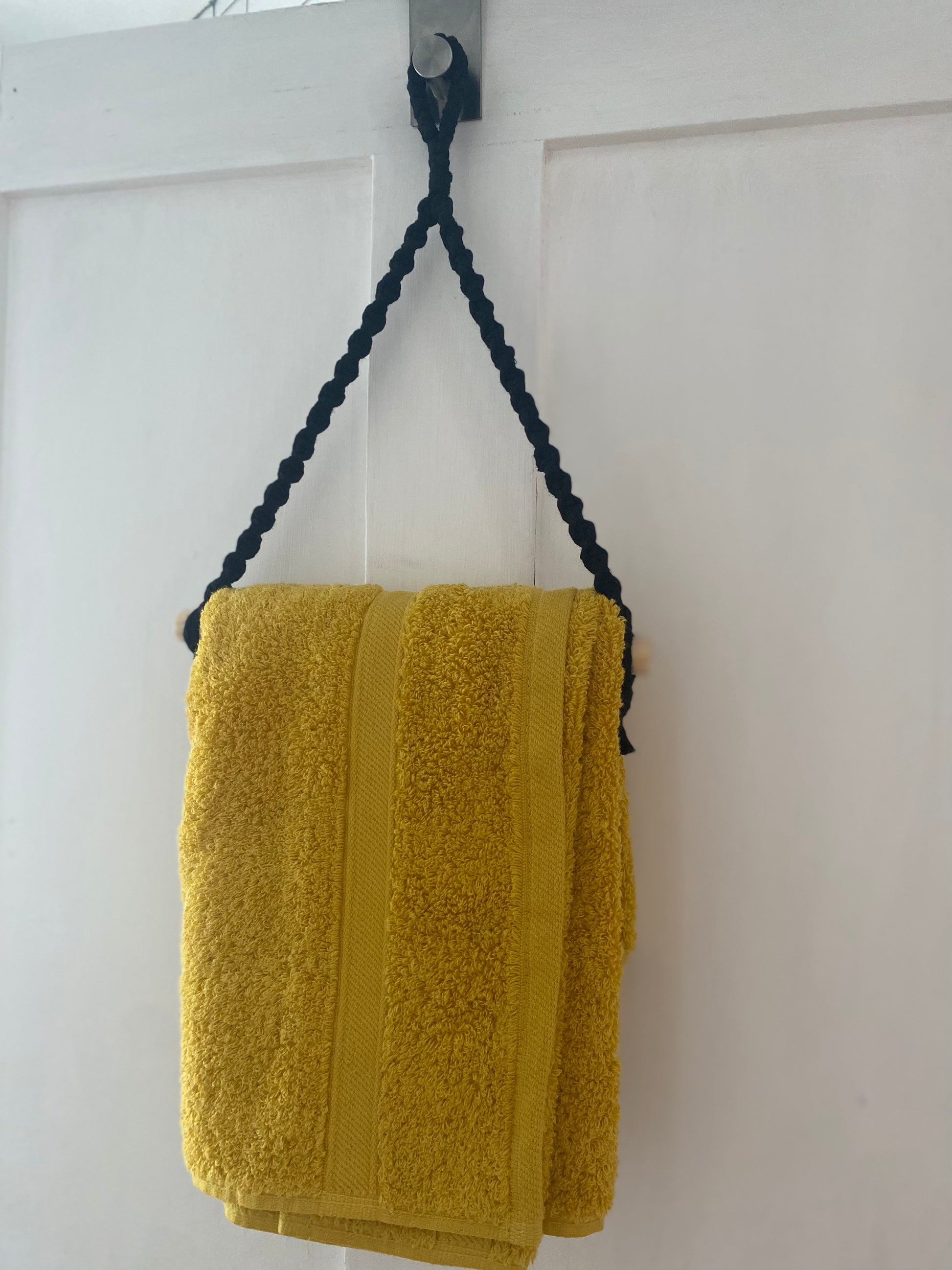 A yellow towel made from sustainable recycled cotton is hanging on a white wooden door by a black braided cord and wooden rod from the Hand towel holder by Macra-Made-With-Love. The towel is neatly folded, with two vertical stripes along its edge. The simple, white-painted door contrasts with the bright eco-friendly towel.