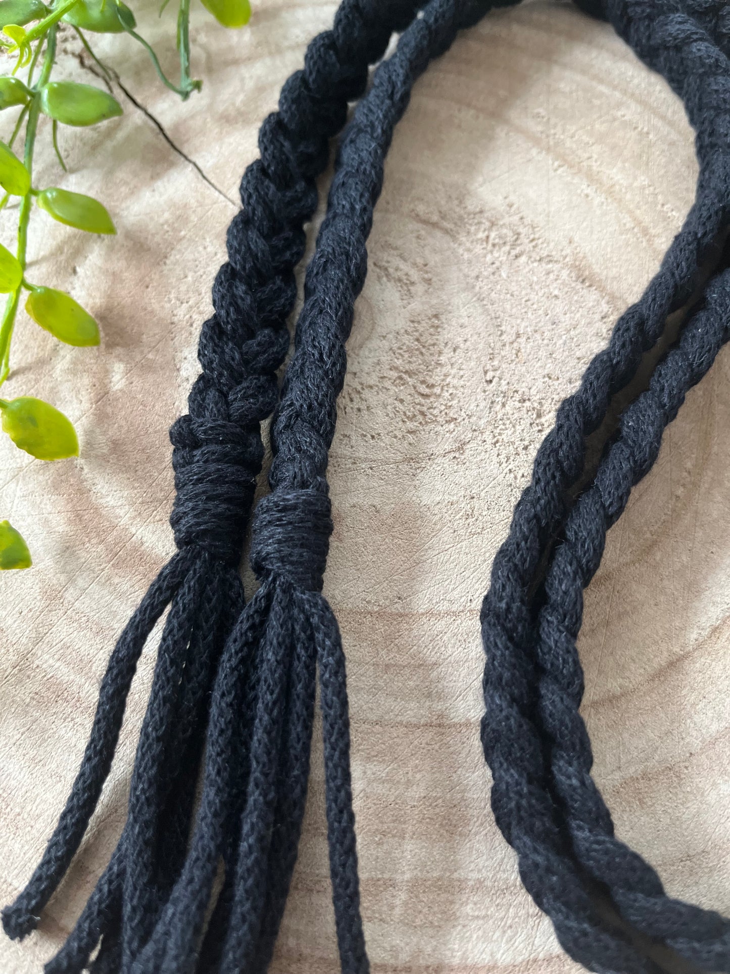 A close-up image of two black braided cords lying on a wooden surface. The ends of the cords are untied into loose strands, showcasing their intricate craftsmanship. A green leafy plant is partially visible along the left edge, complementing this sustainable handmade Braided macrame belt from Macra-Made-With-Love.