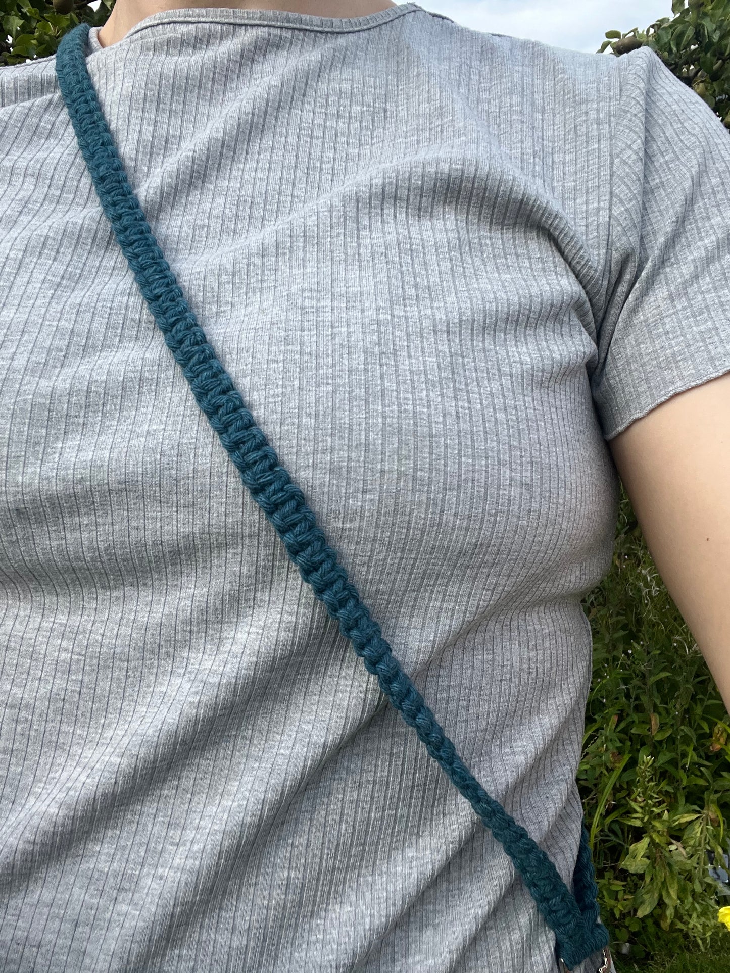 Close-up of a person wearing a light gray ribbed t-shirt, showcasing the Macra-Made-With-Love Recycled Cotton Adjustable Crossbody Phone Strap with Universal Tether, featuring an eco-friendly green knitted design made from recycled cotton, diagonally across their chest. The lush greenery in the background indicates an outdoor setting.