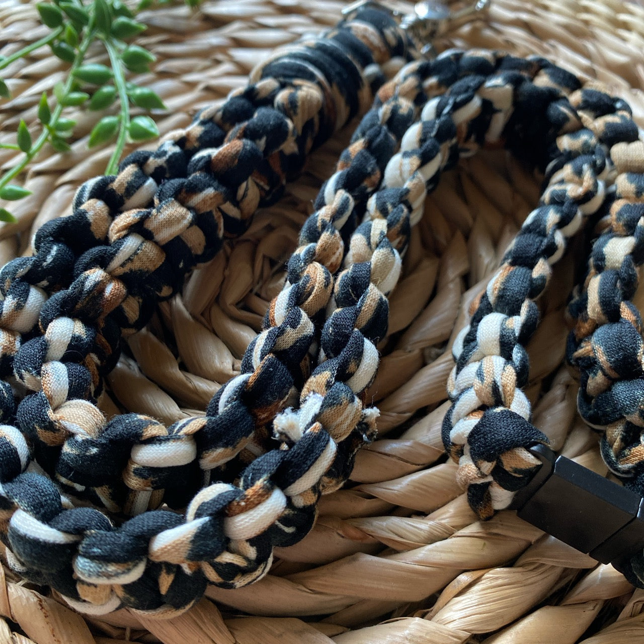 A close-up image of a handmade Eco friendly lanyard badge holder - 34” woven from fabric strips in black, white, and brown colors. It is coiled on a woven basket, with a small green plant in the background. The texture and pattern of this Macra-Made-With-Love eco-friendly lanyard are prominently visible.