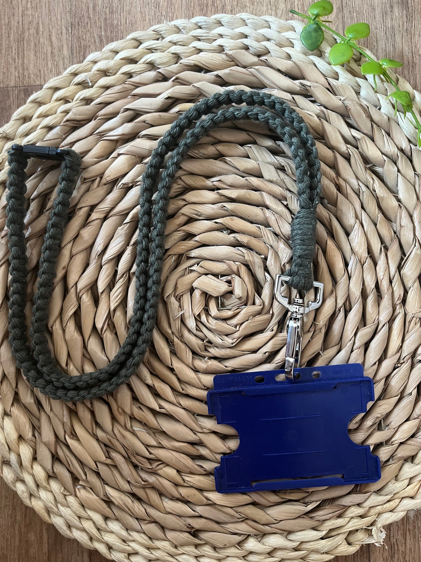 A blue plastic ID badge holder attached to the Eco friendly lanyard badge holder - 34” by Macra-Made-With-Love is placed on a woven circular mat. A small green plant is partially visible in the top right corner of the mat, which rests on a wooden surface.