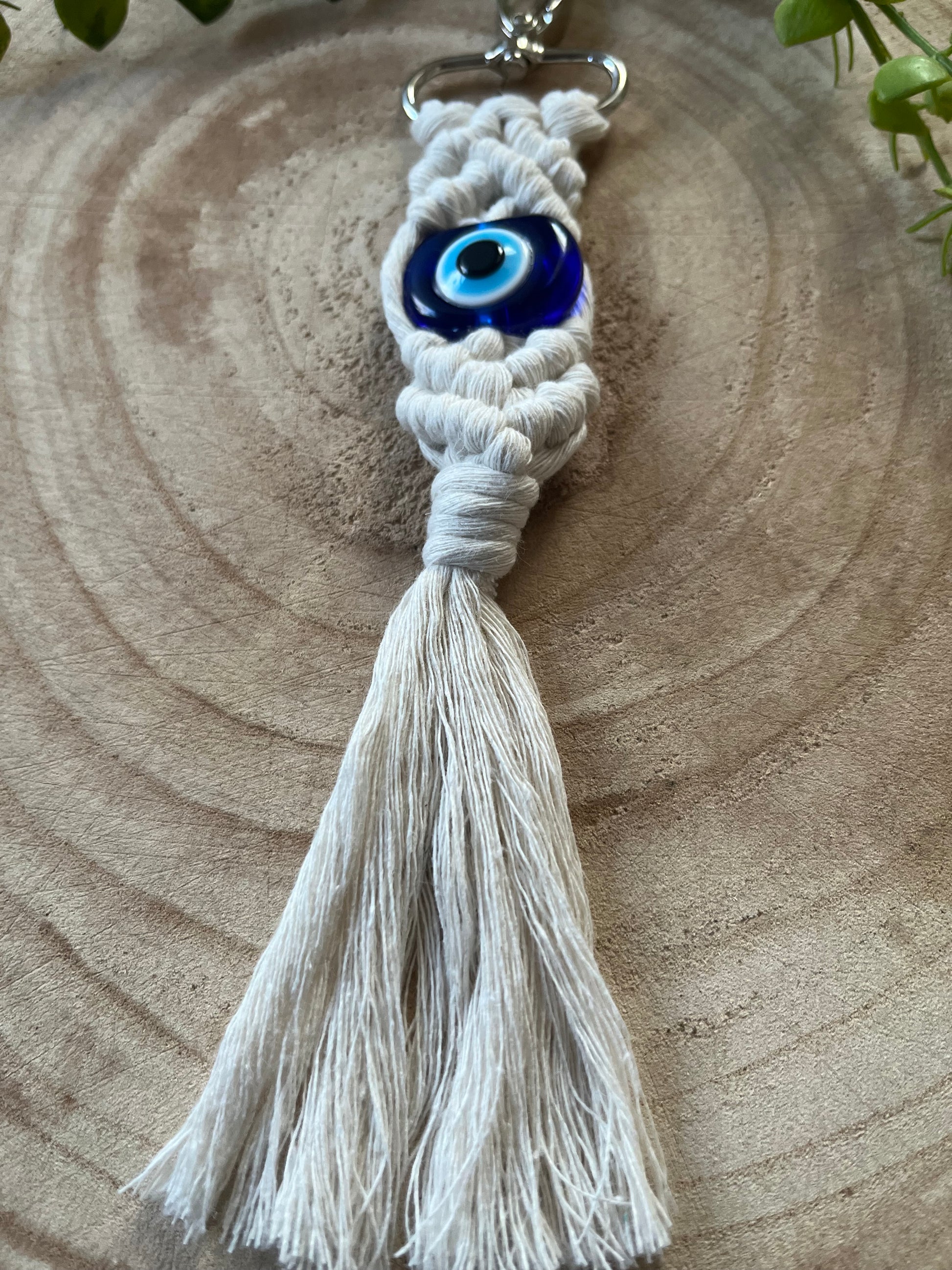 A close-up of an Evil Eye Keychain featuring intricate knots and a large blue evil eye bead at the center. This handmade Evil eye keychain from Macra-Made-With-Love has a white tassel at the bottom and is placed on a wooden surface with some green leaves in the background, serving as both a stylish accessory and a protection talisman.