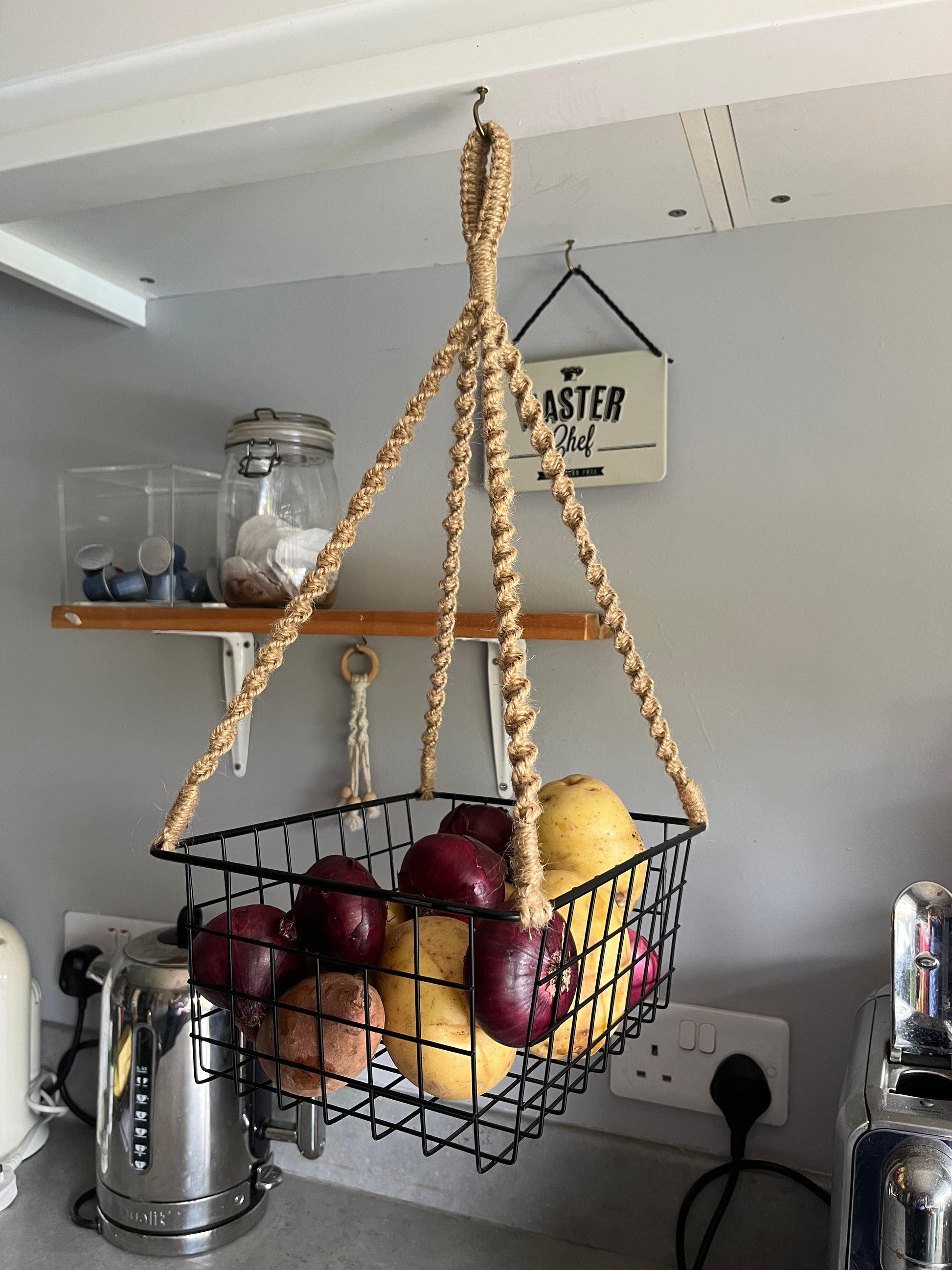 Black metal basket hanging from a jute rope, holding red onions and white potatoes. 
