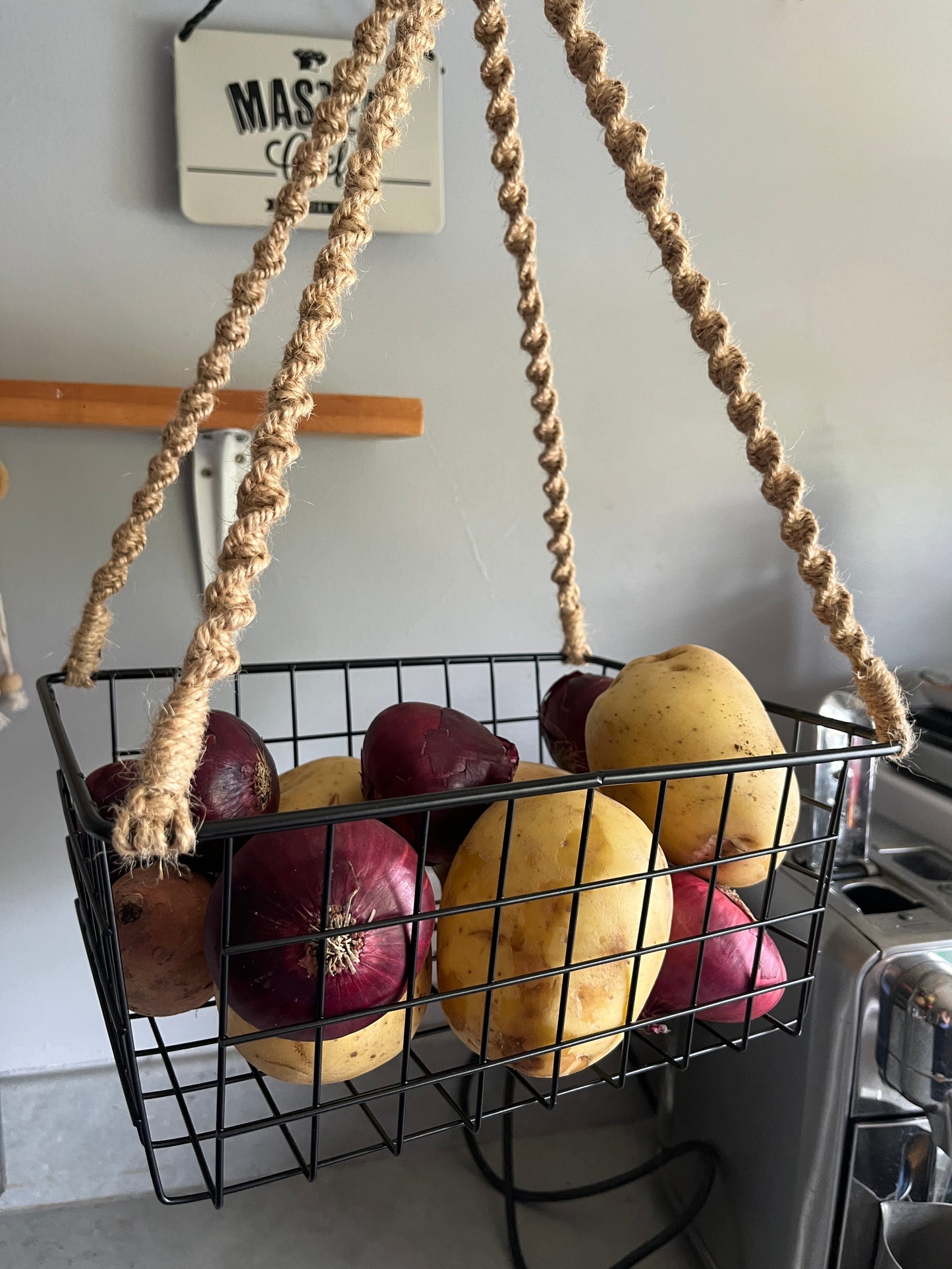 Wire basket with potatoes and onions hanging from a jute rope against a grey kitchen wall.