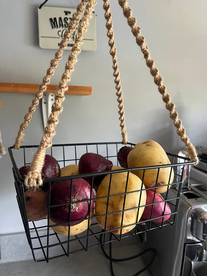 Wire basket with potatoes and onions hanging from a jute rope against a grey kitchen wall.