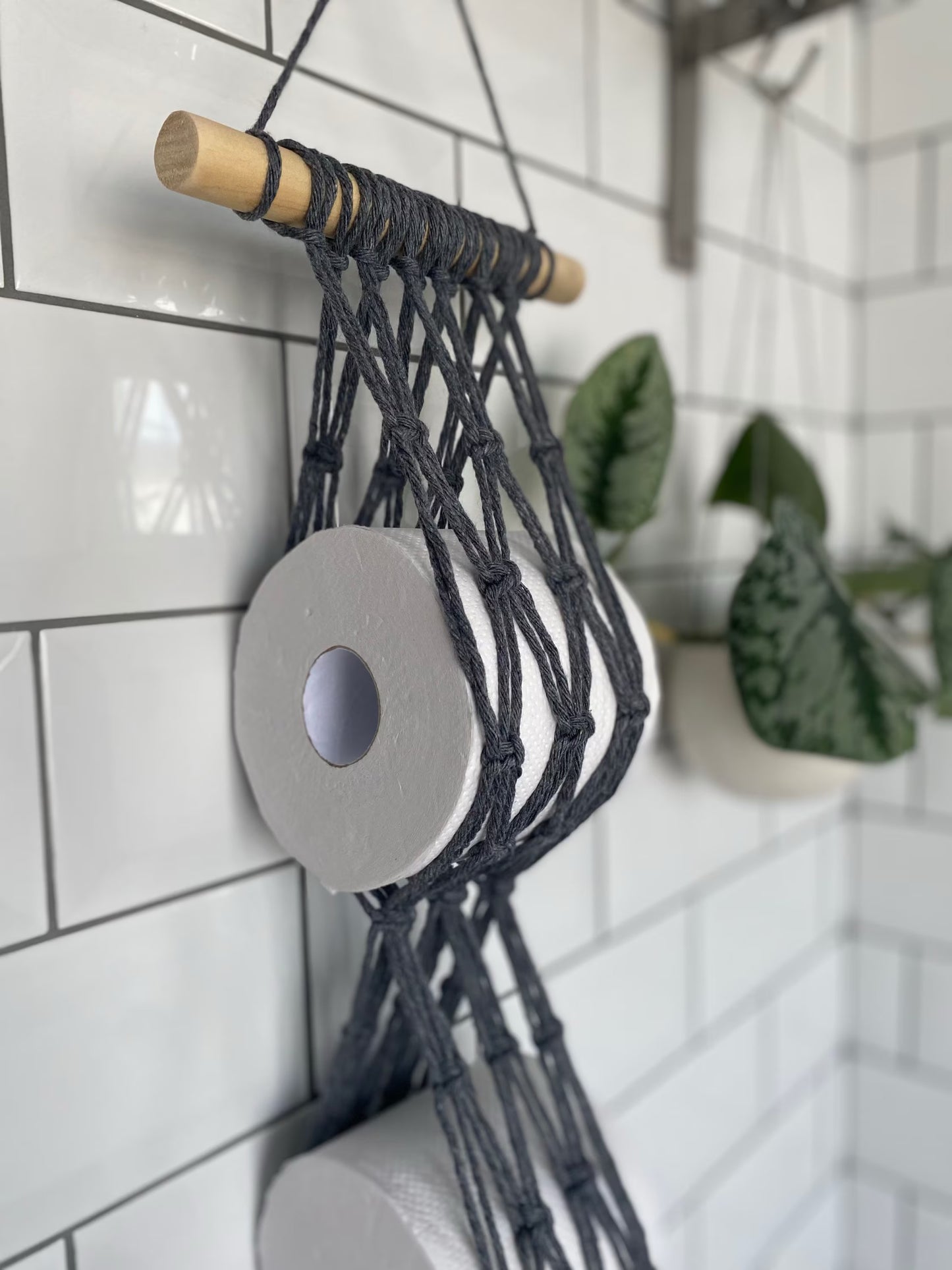 A Macra-Made-With-Love Toilet paper wall mounted storage, an exquisite piece of bathroom decor, hangs on a tiled wall holding two rolls of toilet paper. Made of dark gray, knotted cords attached to a wooden rod, this eco-friendly accessory is complemented by a plant with green leaves in a white pot in the background.