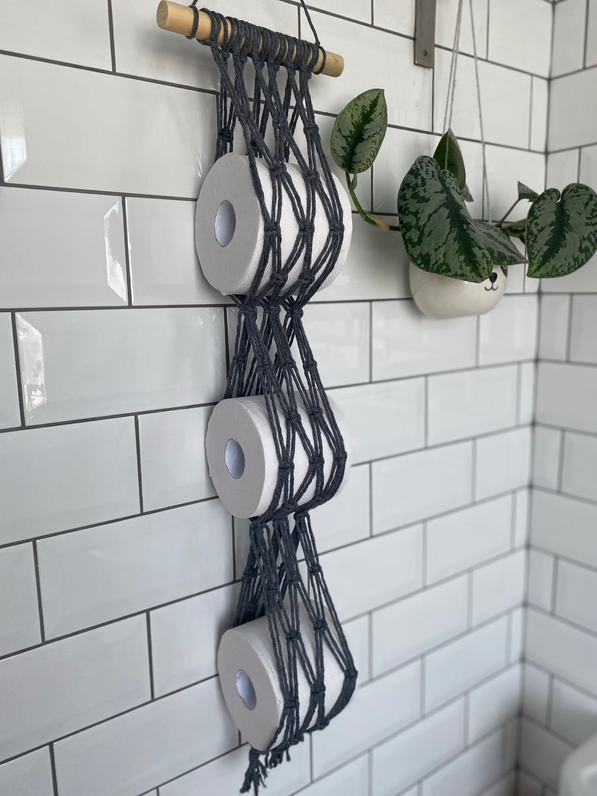 A decorative, eco-friendly Macra-Made-With-Love Toilet paper wall mounted storage hangs from a tiled white wall, holding three rolls vertically. Nearby, a potted plant in a white hanging planter adds a touch of greenery to the minimalist bathroom decor.