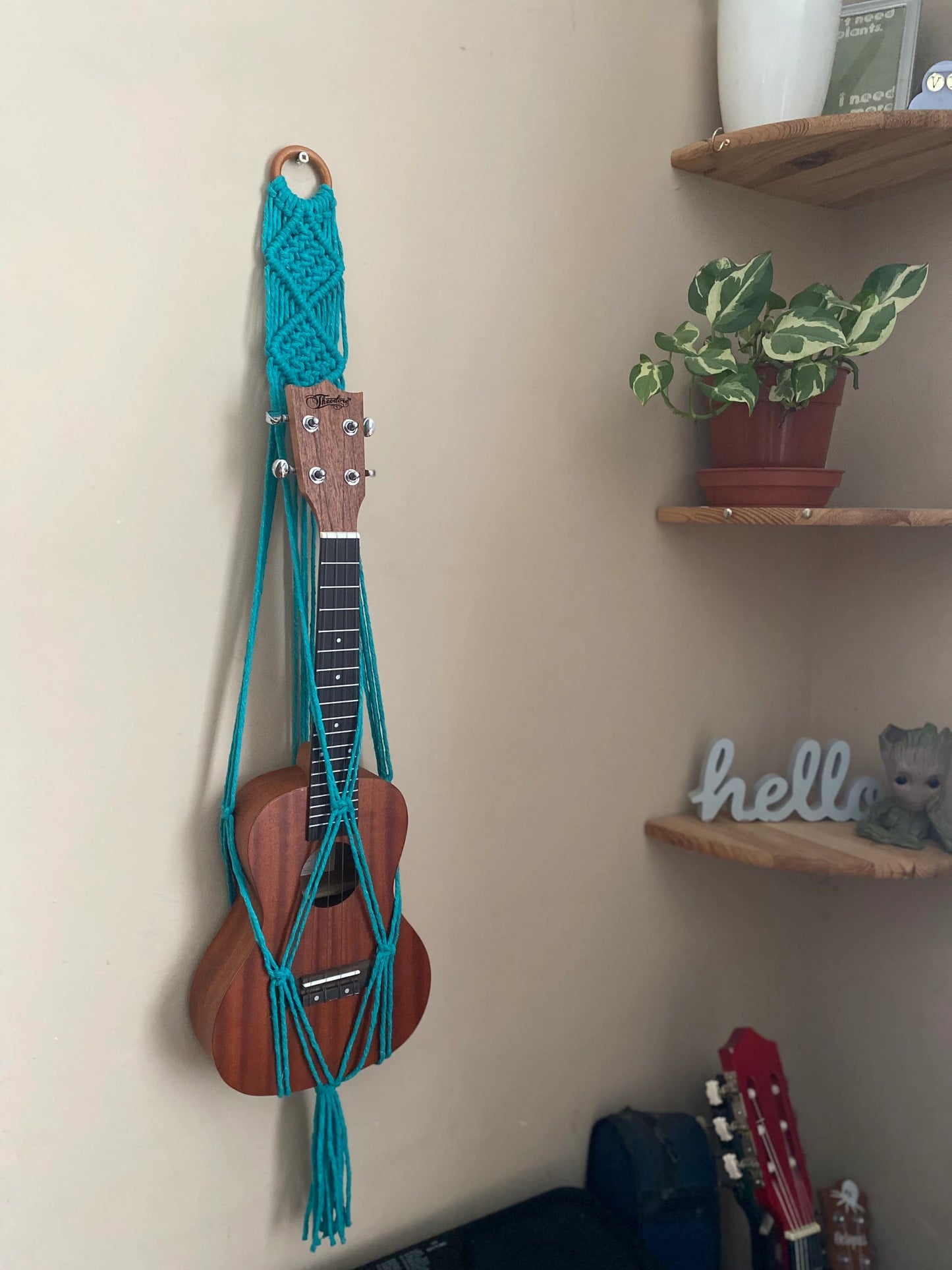 A dark brown ukulele sits inside of a handmade macrame wall hanging ukulele hanger, which is made with turquoise blue recycled cotton, and a repurposed wooden hanging ring. A set of corner shelves is to the right of the ukulele, with a small plant on them. 