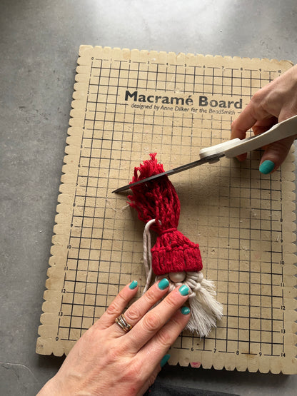 A person's hands are shown crafting a macrame Santa decoration on a macrame board, with cotton cords and wooden bead.