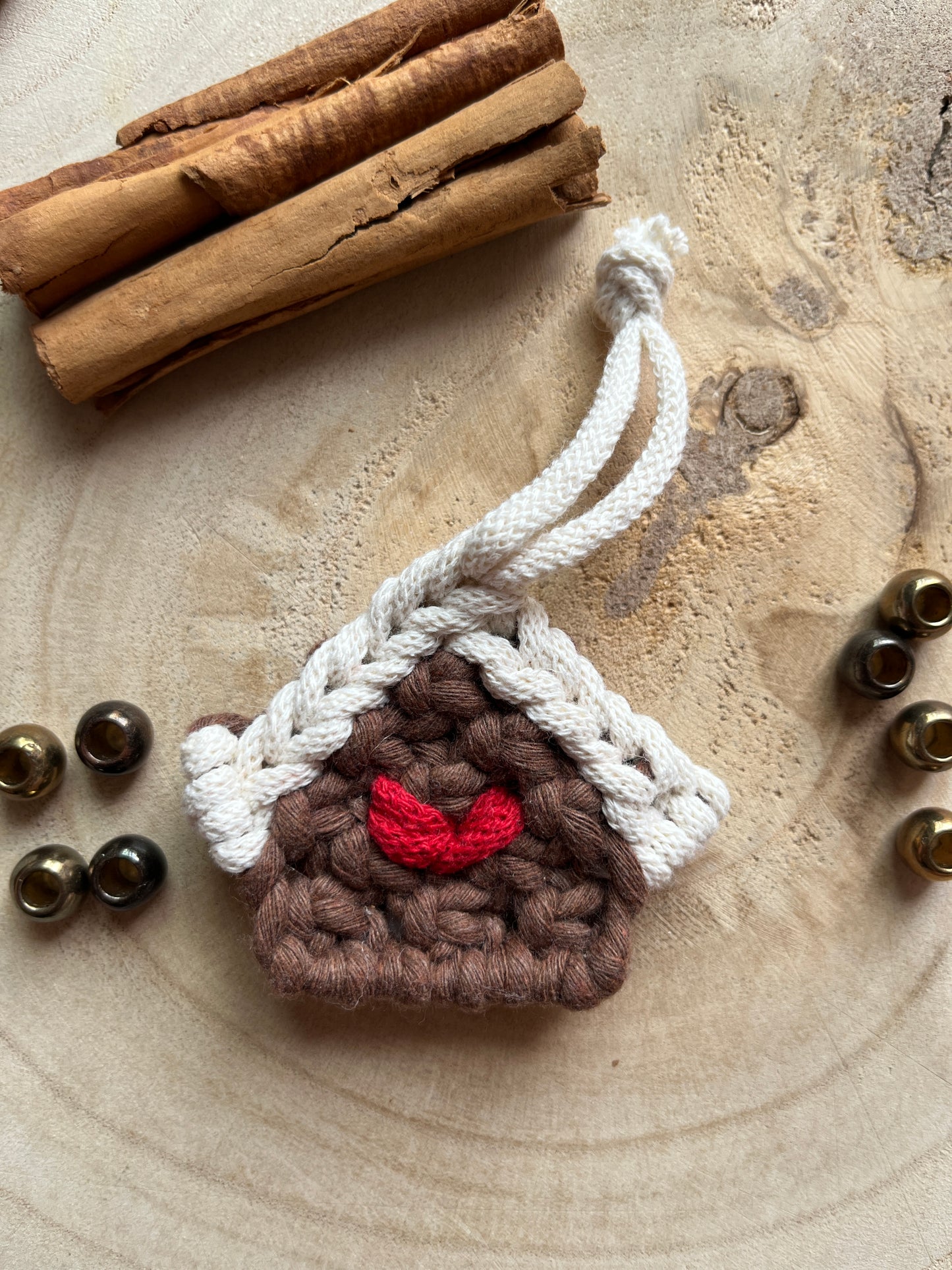 A small macramé decoration in the shape of a gingerbread house, made with brown and white yarn with a red heart detail in the middle, styled on a light wood surface with cinnamon sticks and metal beads. 