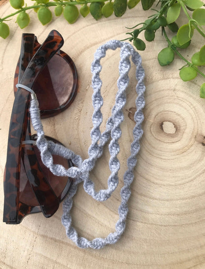 Sunglasses with a light grey strap on a wooden surface with green leaves.