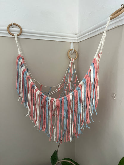 Macrame hammock, made with pink, blue and white yarns and three large wooden rings, hanging in the corner of a room with beige painted walls. The hammock is empty. 
