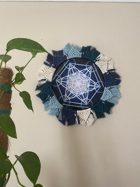 A macrame mandala frame knotted with blue and white yarn, surrounds a Metatron’s Cube geometric print. Hung on a beige wall next to a tall houseplant. 