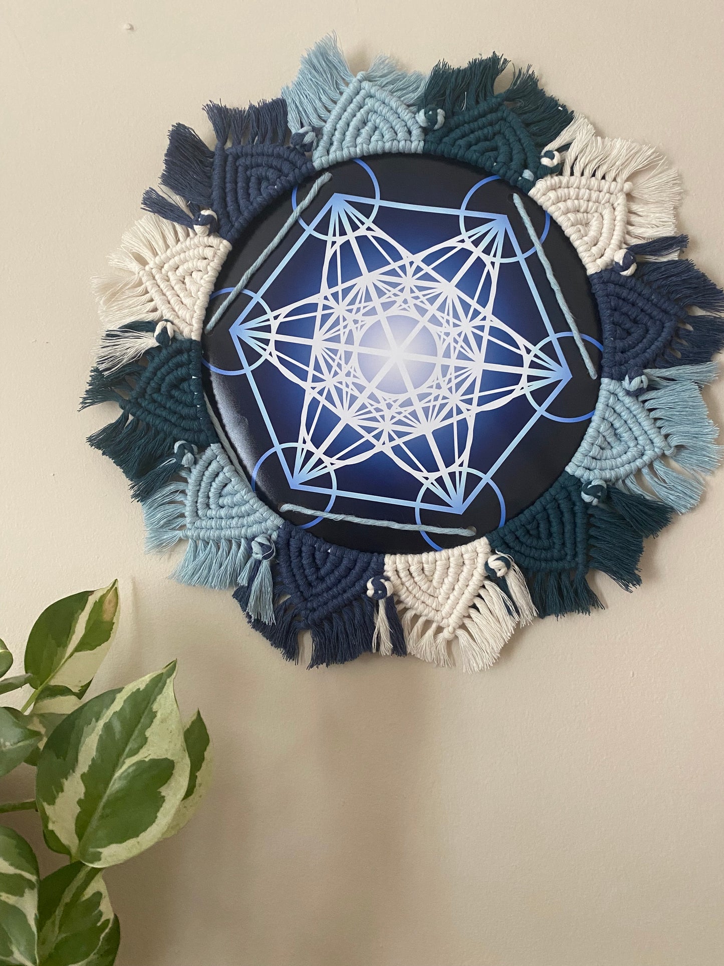 A macrame mandala frame knotted with blue and white yarn, surrounds a Metatron’s Cube geometric print. Hung on a beige wall next to a small houseplant. 