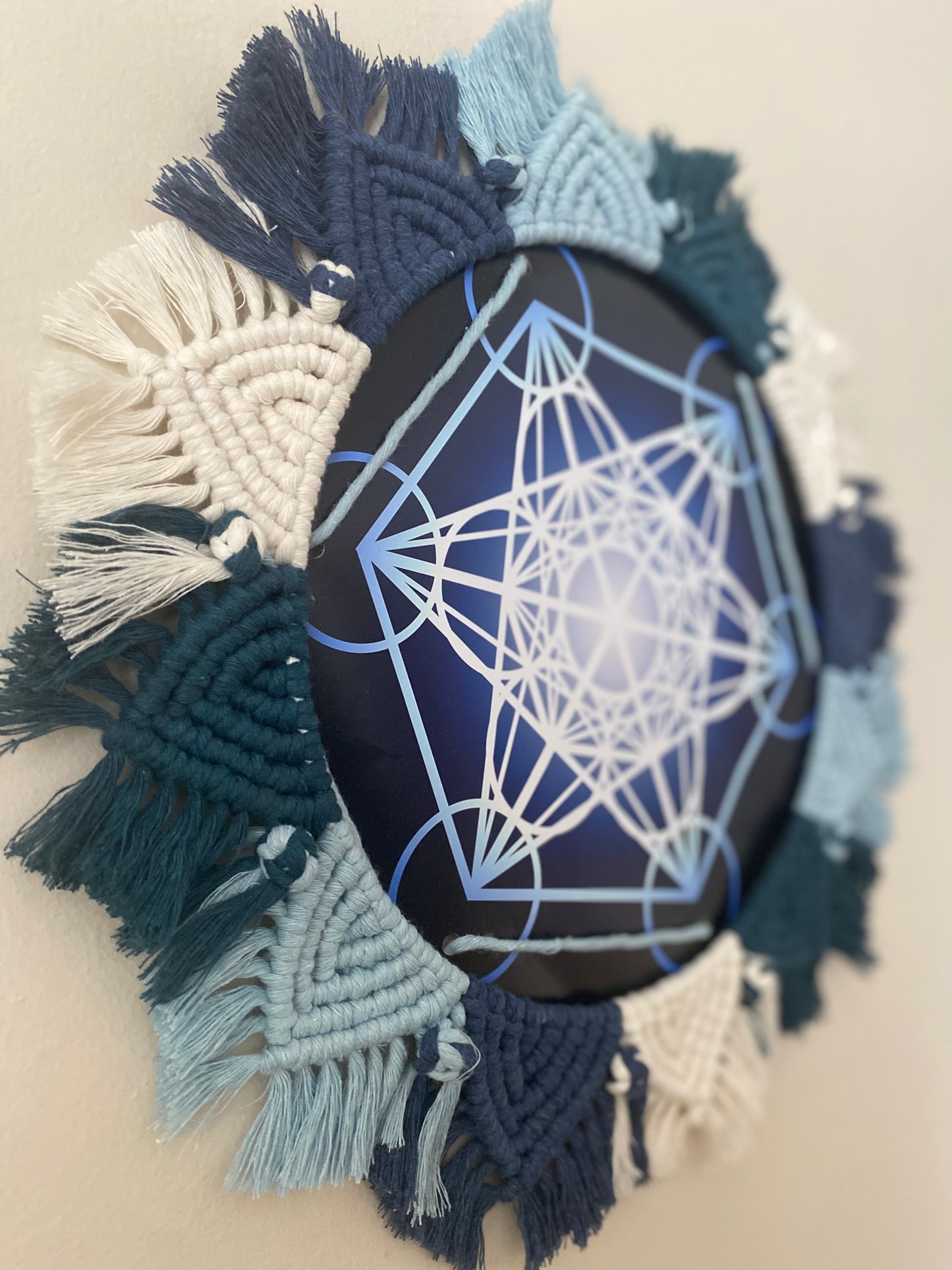 A close up focused image of a macrame mandala frame knotted with blue and white yarn, surrounding a Metatron’s Cube geometric print. 