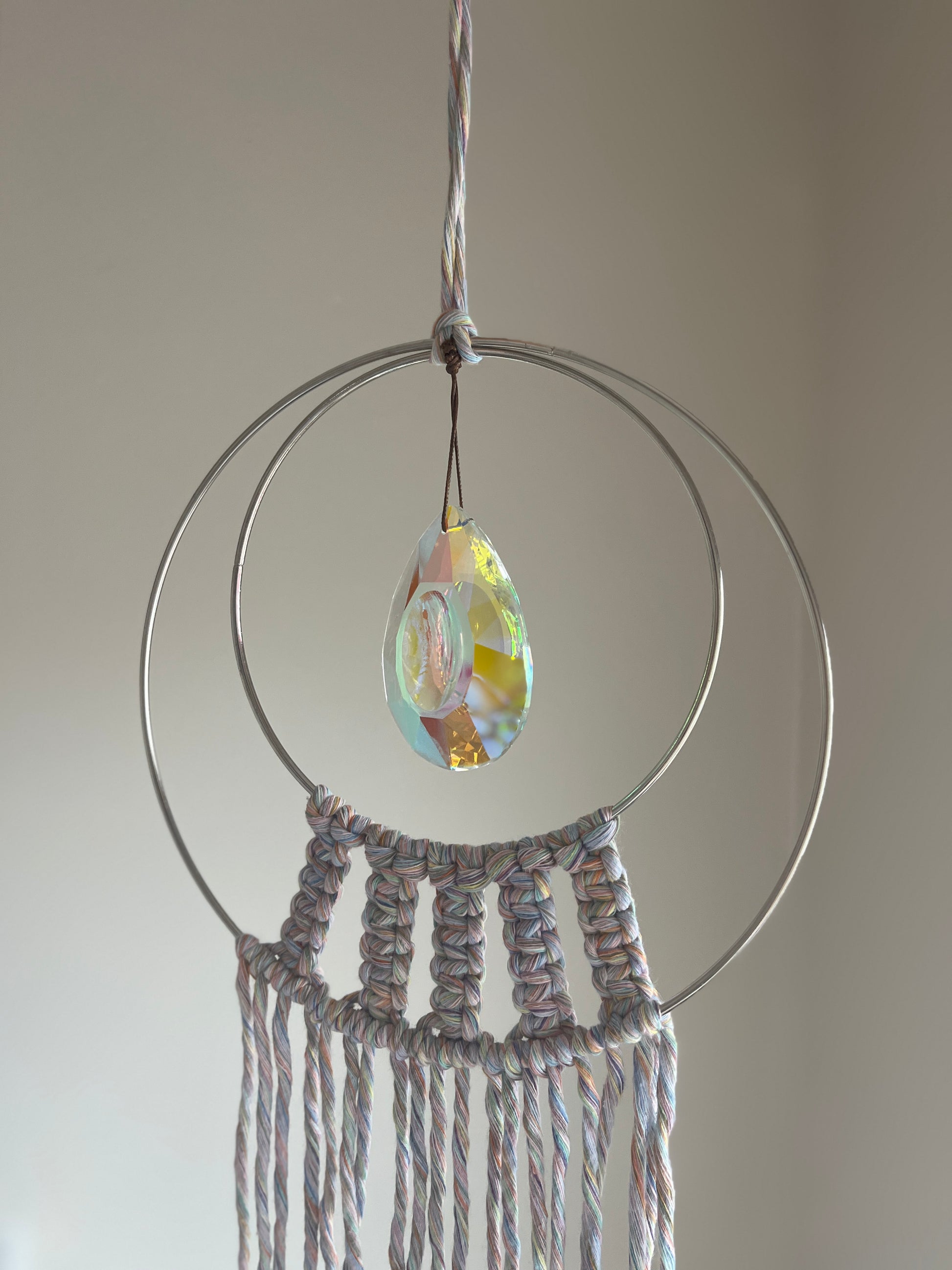 Handmade macrame Suncatcher made with pastel coloured recycled yarn, on two metal hoops, with a large hanging crystal in the centre, shown hanging in front of a white wall. 