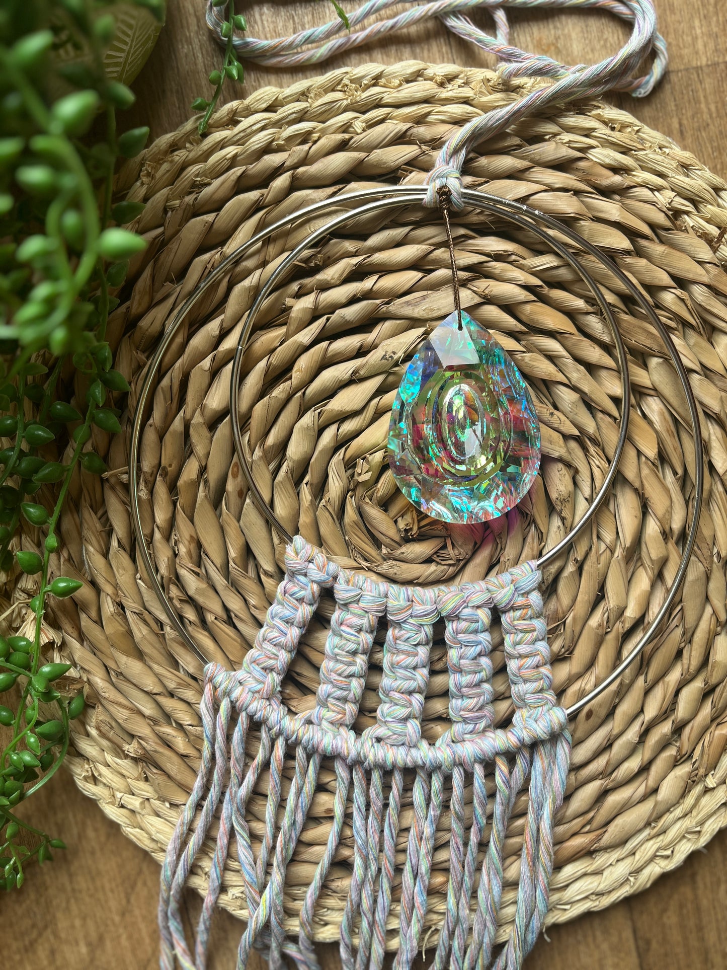 Handmade macrame Suncatcher made with pastel coloured recycled yarn, on two metal hoops, with a large hanging crystal in the centre, laid on a wicker surface 