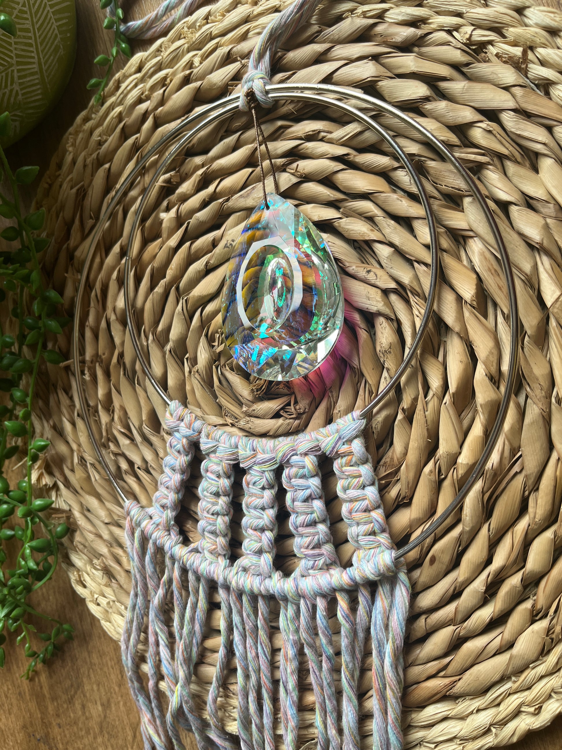Angled close up of a Handmade macrame Suncatcher made with pastel coloured recycled yarn, on two metal hoops, with a large hanging crystal in the centre. 