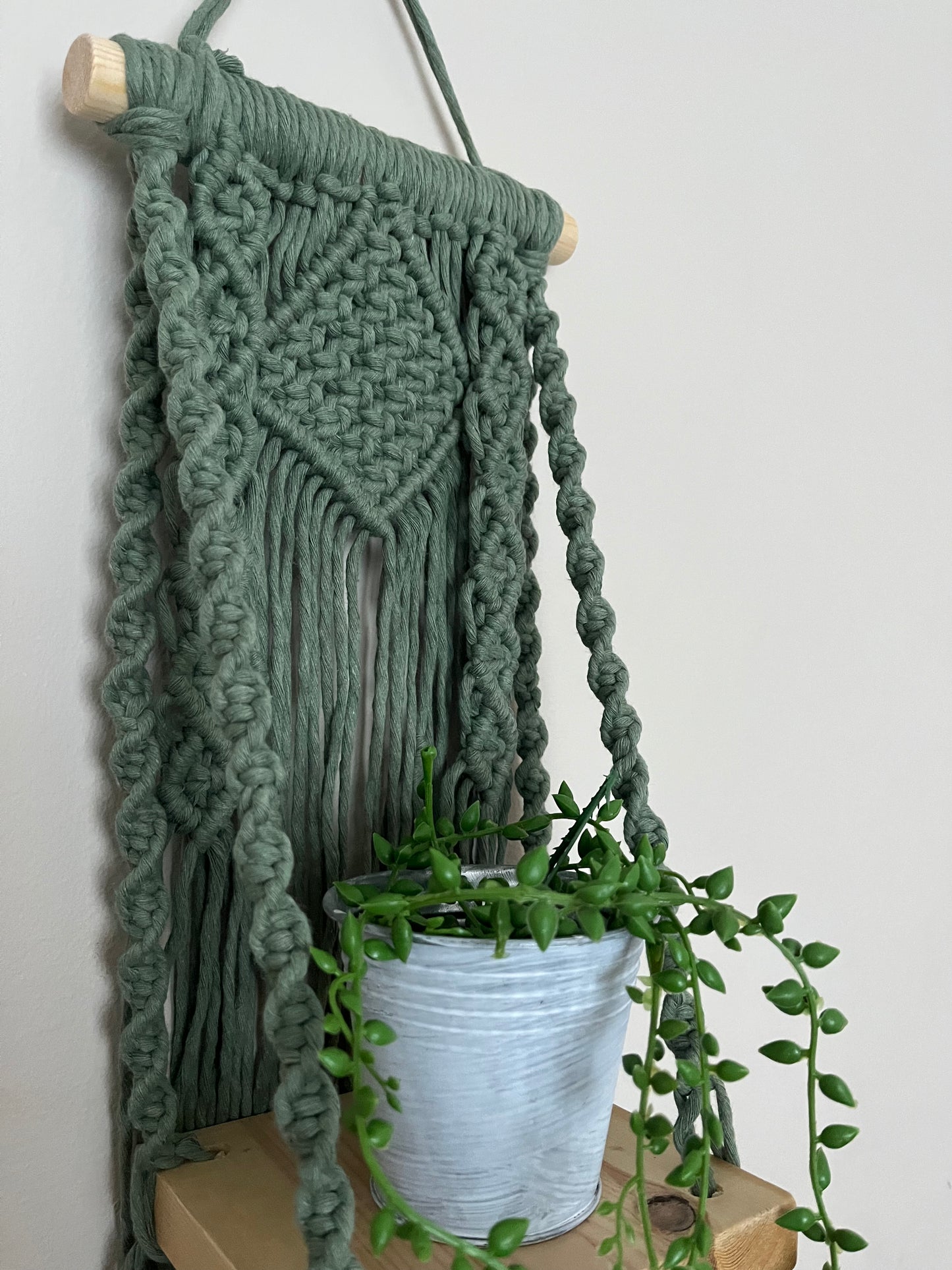 Handmade macrame boho style shelf, made with sage green recycled cotton on a solid wooden dowel, with a small square shelf. The shelf has an intricately knotted pattern, and displays a small artificial plant. 