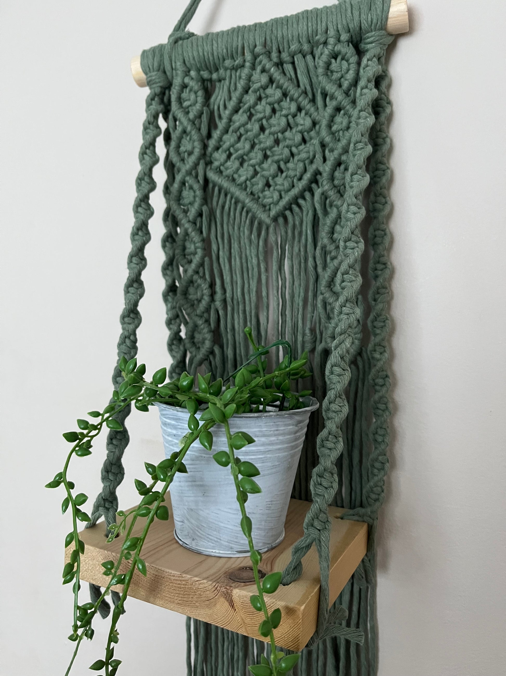 Handmade macrame boho style shelf, made with sage green recycled cotton on a solid wooden dowel, with a small square shelf. The shelf has an intricately knotted pattern, and displays a small artificial plant. 